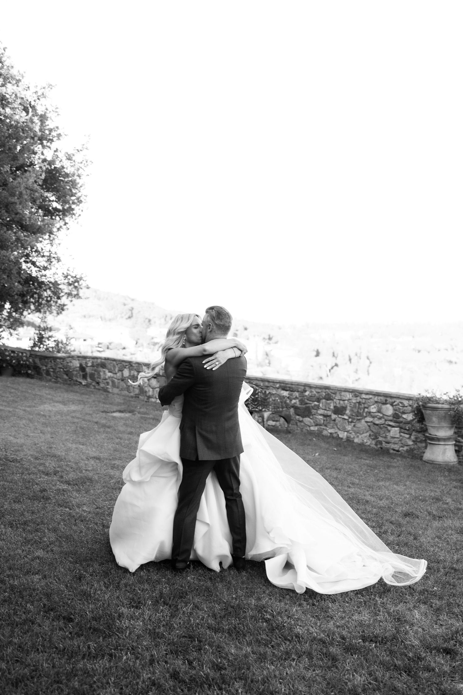 A black and white wedding photograph showing a couple embracing on a grassy lawn with stone walls in the background.