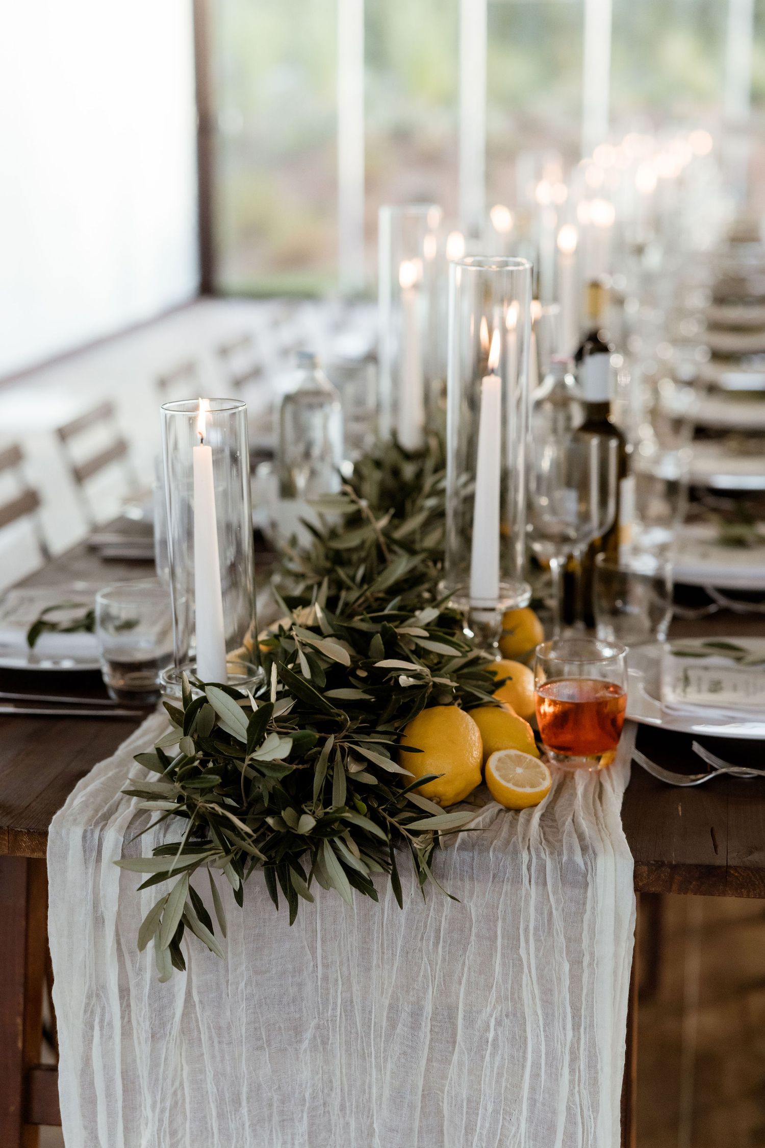 Elegant table setting with white linens, tall glass candleholders, olive branches, and citrus fruit decorations.