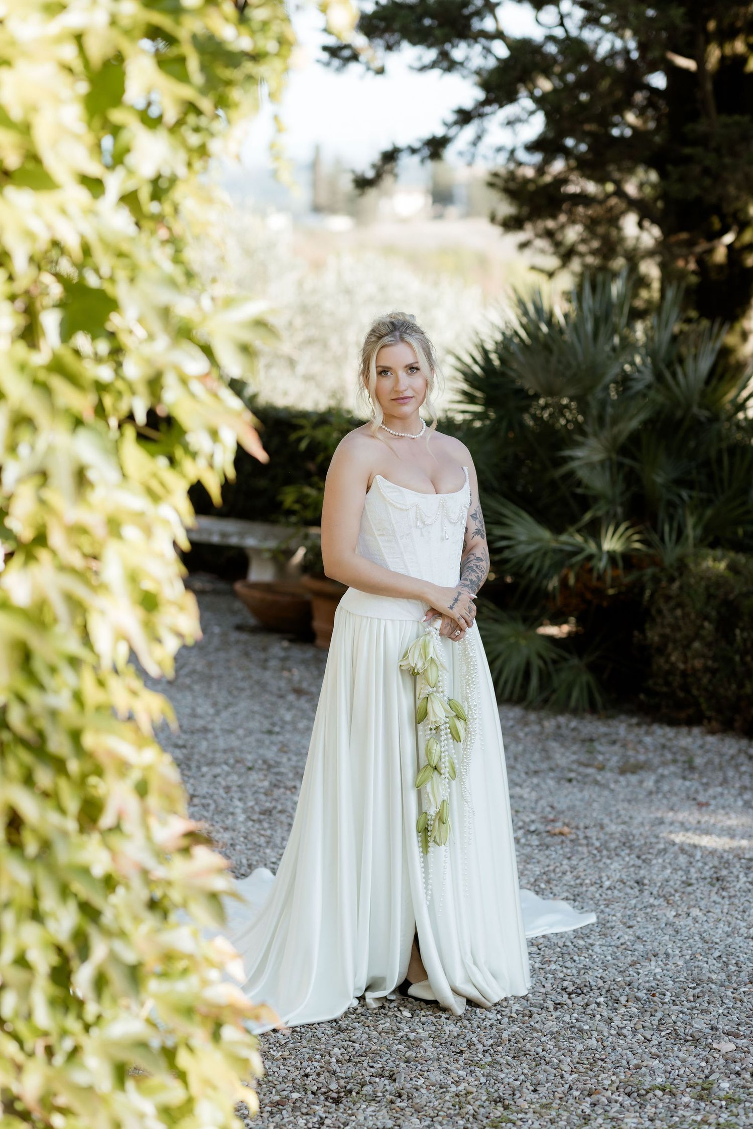 Bride in a flowing white empire waist gown poses gracefully on a gravel path surrounded by lush garden foliage.