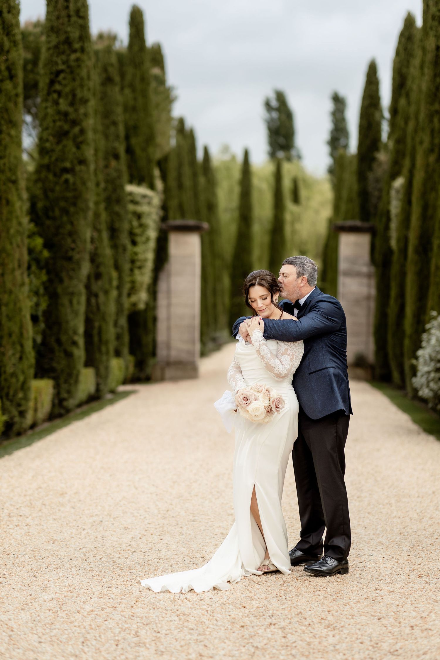 A romantic moment captured in a cypress tree-lined pathway at an elegant outdoor venue.