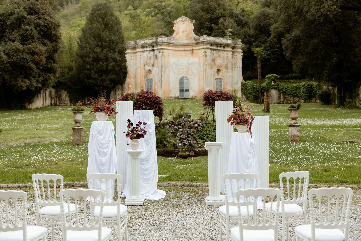 Elegant garden wedding ceremony setup with white chairs and floral arrangements in front of a historic stone pavilion.
