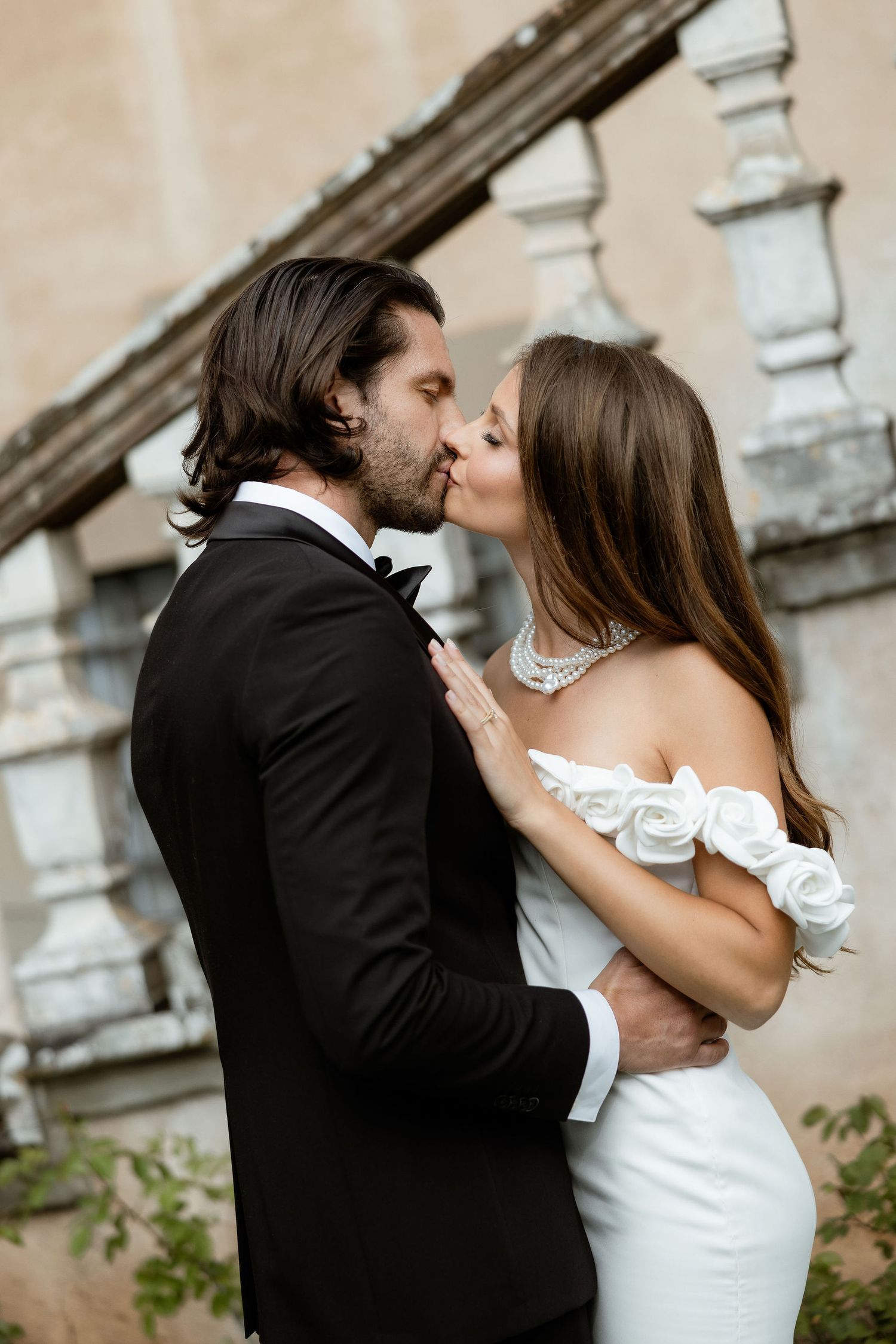 Romantic couple in elegant wedding attire share an intimate kiss in front of ornate stone pillars.