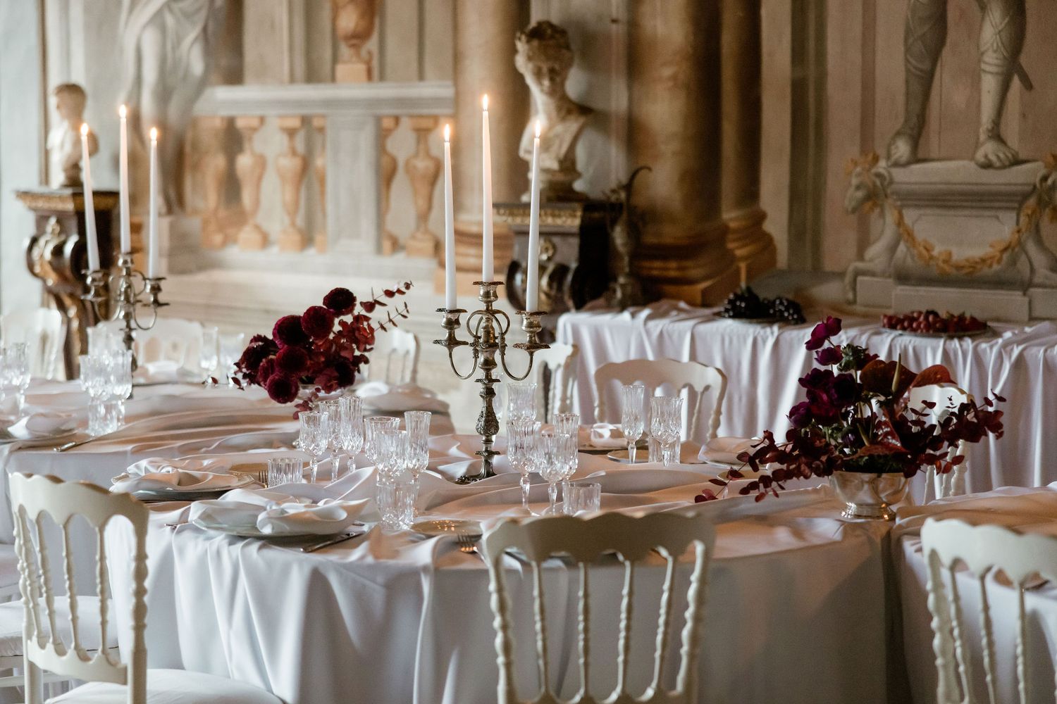 Elegant wedding reception tables with white linens, crystal glassware, and burgundy floral centerpieces in a luxurious venue.