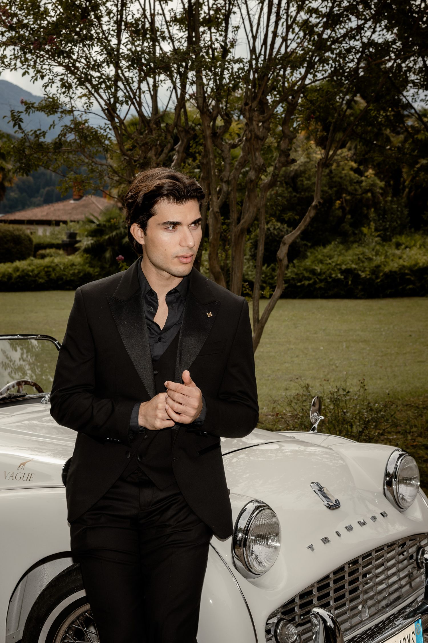 Classic Triumph car poses with fashionable gentleman in black suit against scenic mountain backdrop.