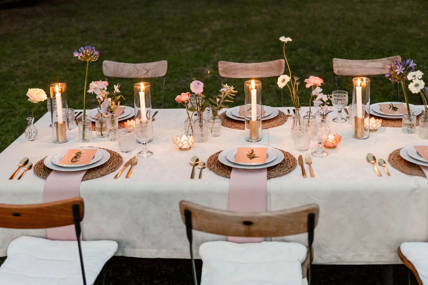 A beautifully set wedding table with white linens, wooden chairs, and gold accents in an outdoor setting.