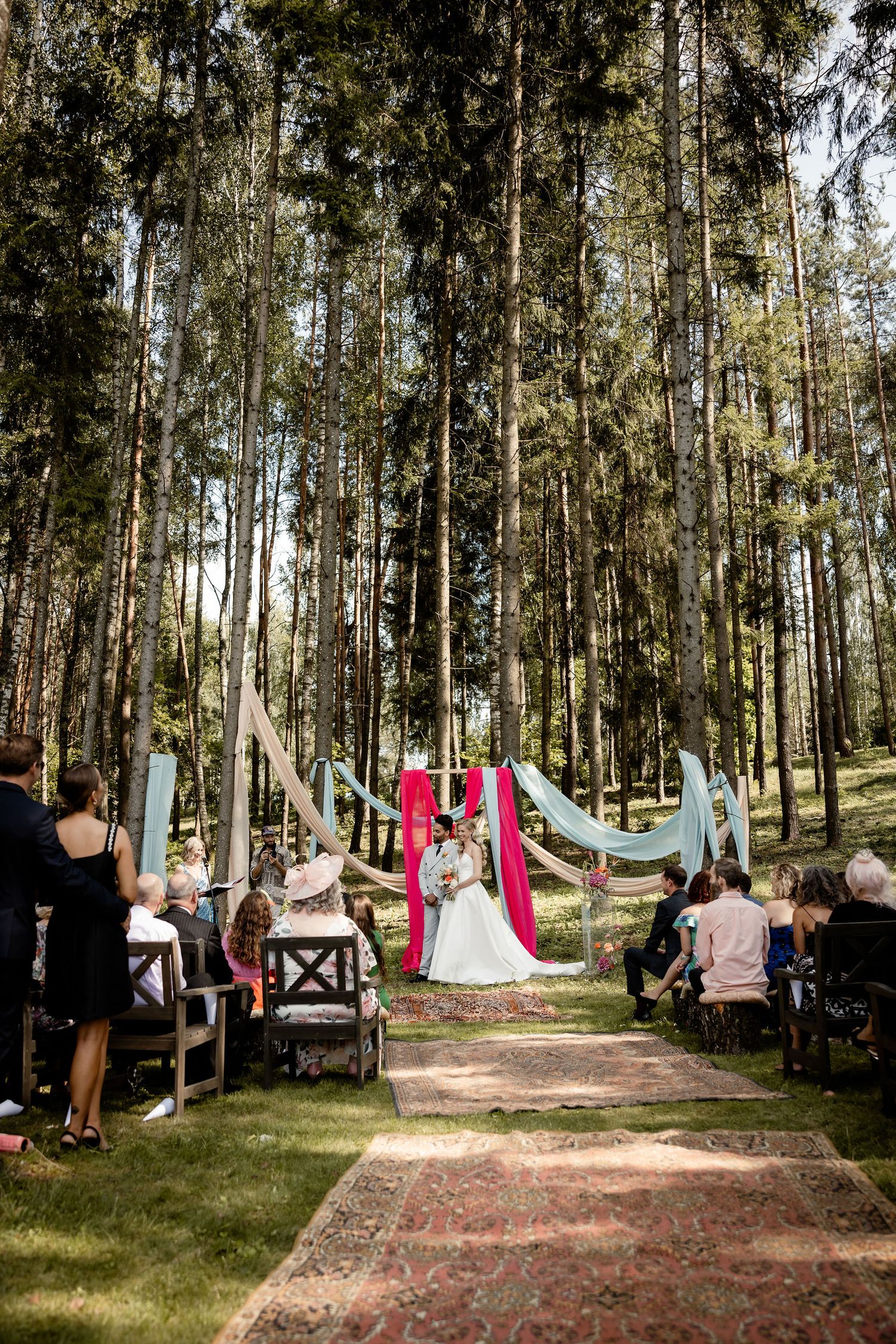 An outdoor forest wedding ceremony setup with red and blue drapes hanging between tall trees.