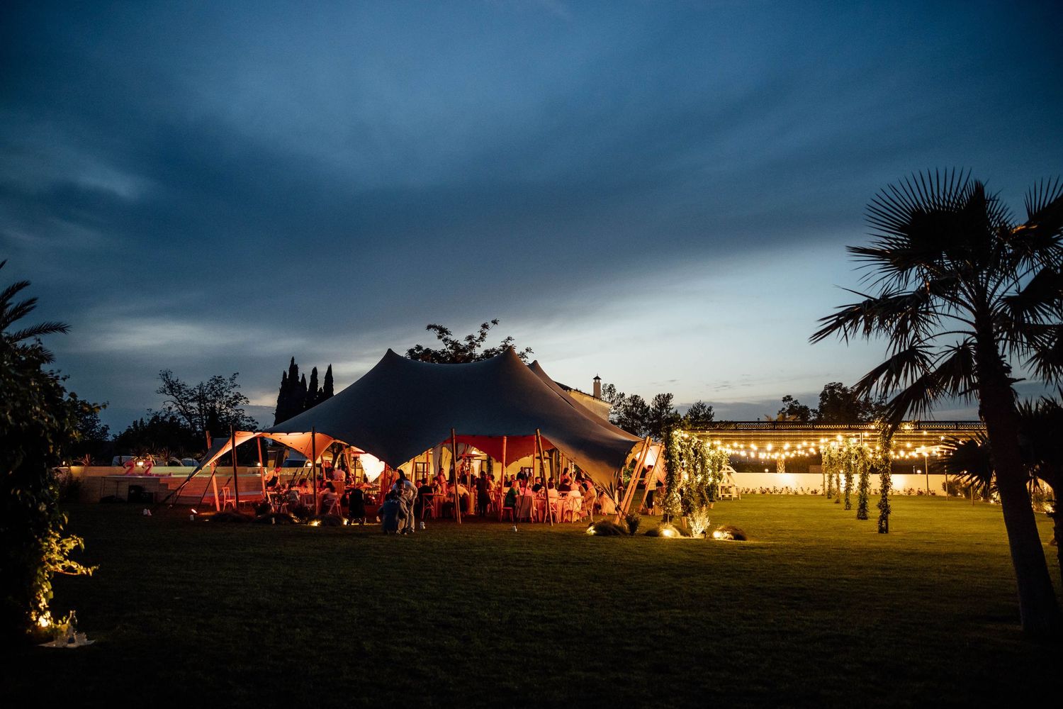 Illuminated wedding tents glowing at dusk with palm trees silhouetted against a moody sky.
