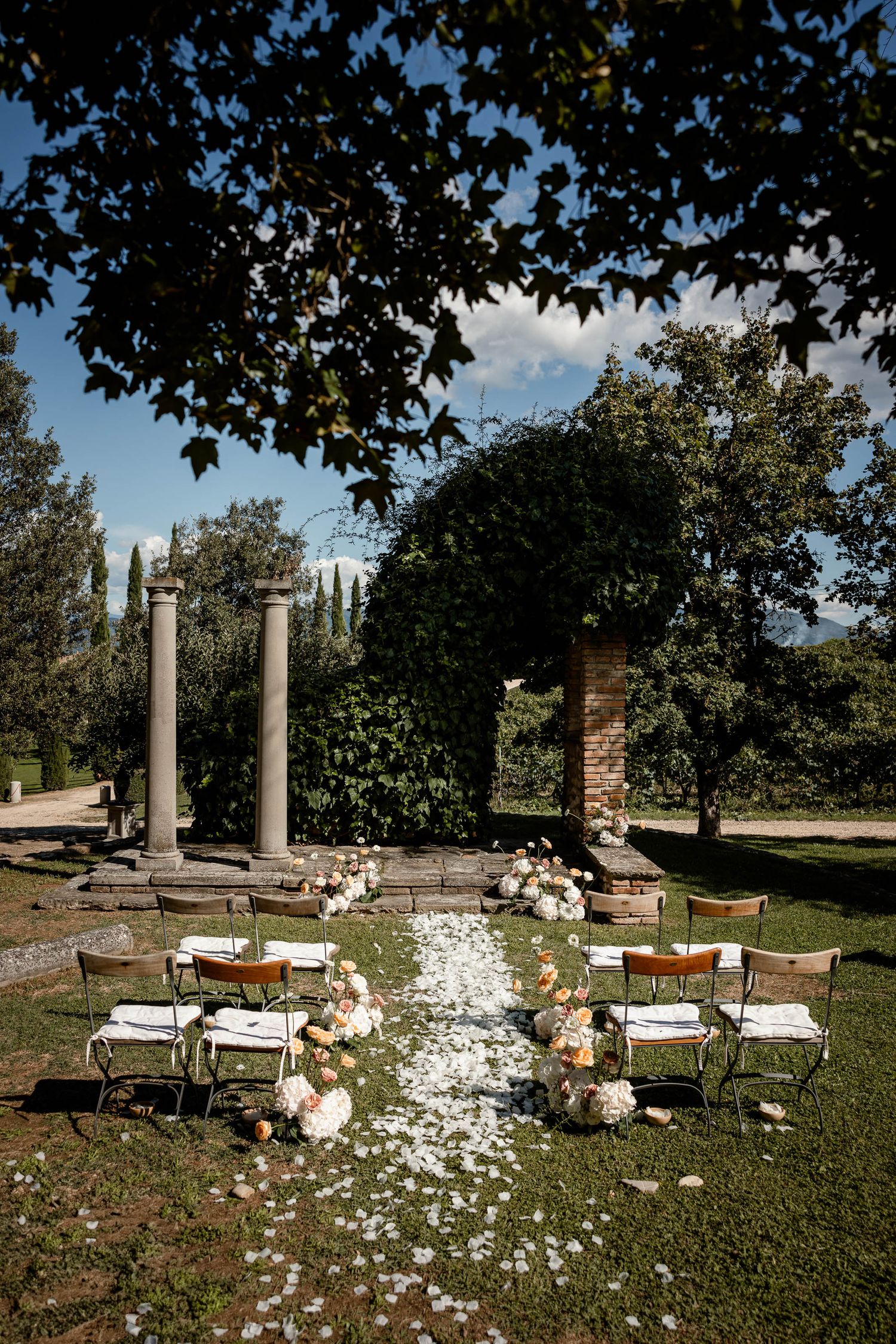 Outdoor wedding ceremony setup with stone columns and flower petals scattered along the aisle.