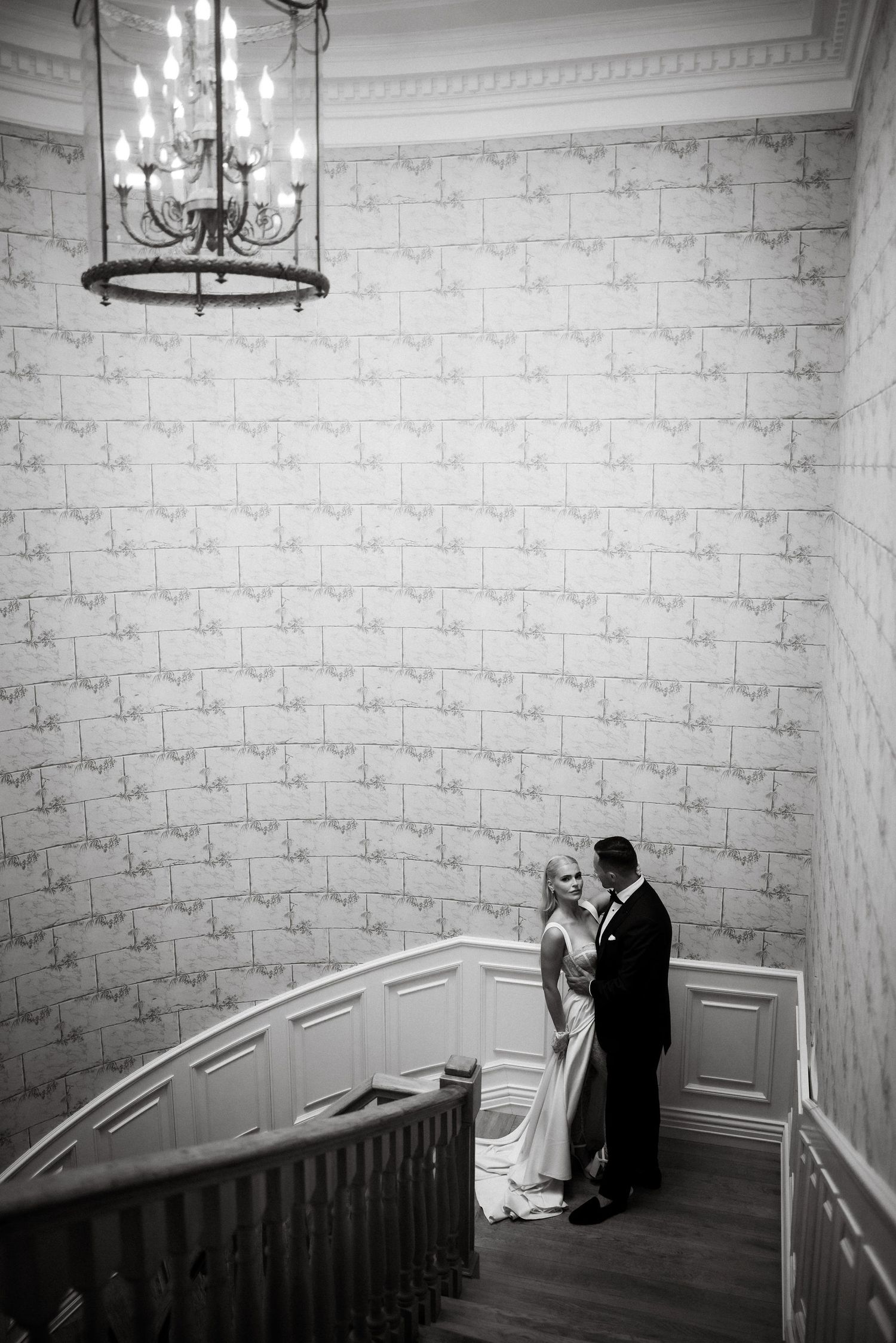 A black and white wedding photo captures a couple's intimate moment on a curved staircase.