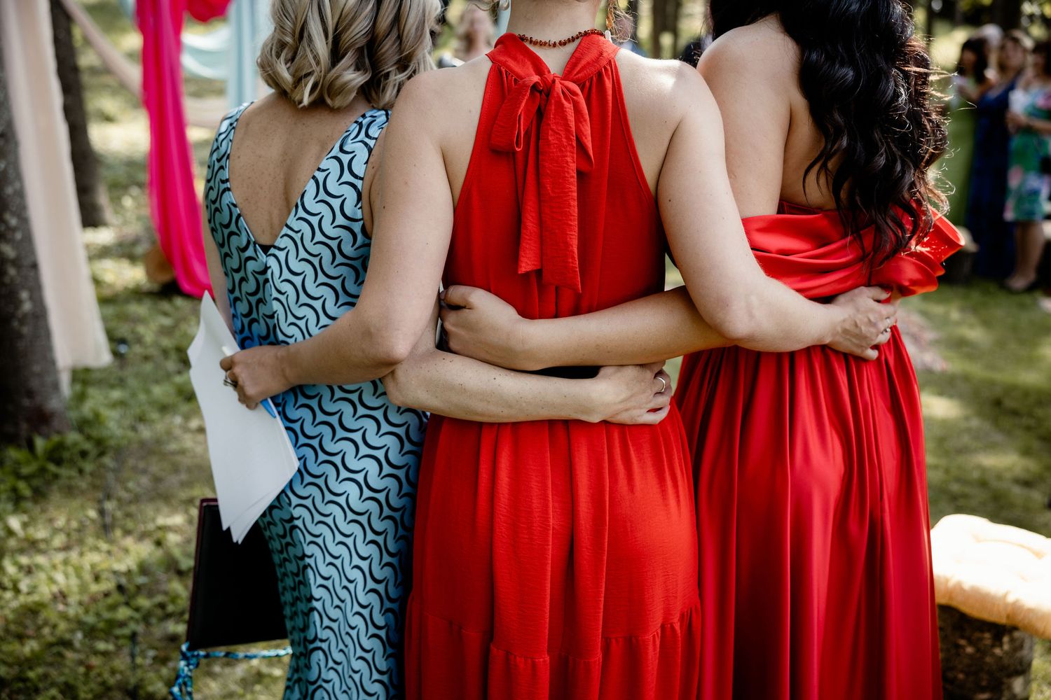 Three women in formal dresses, two wearing vibrant red and one in a patterned blue, stand close together at an outdoor event.