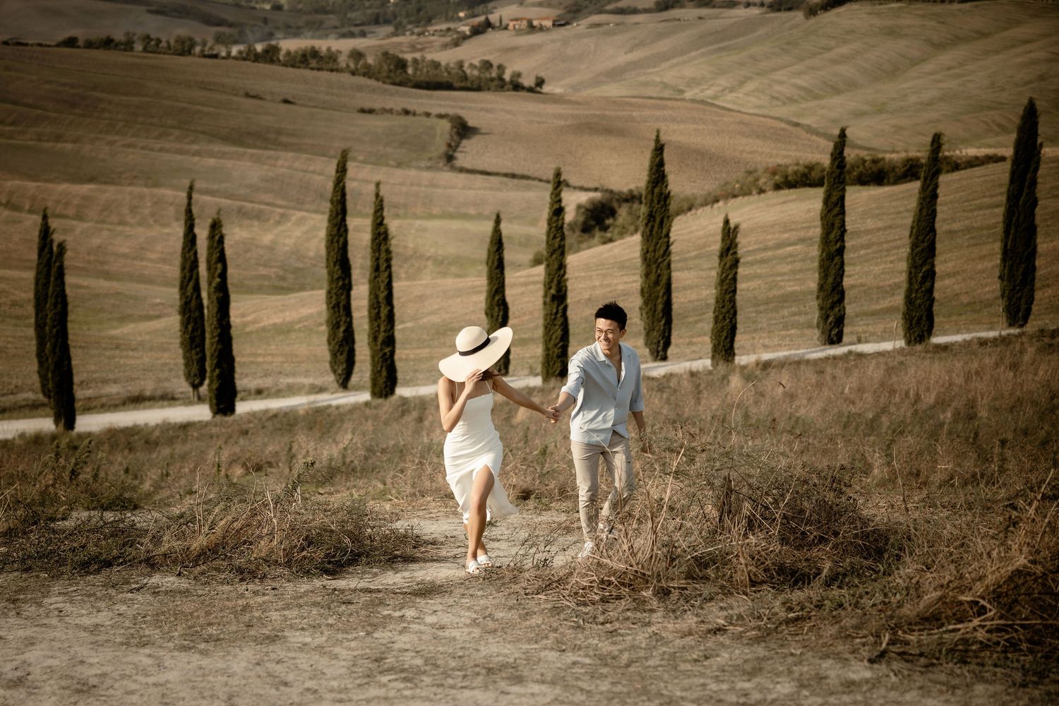 A couple runs playfully down a Tuscan hillside path with cypress trees in the background.