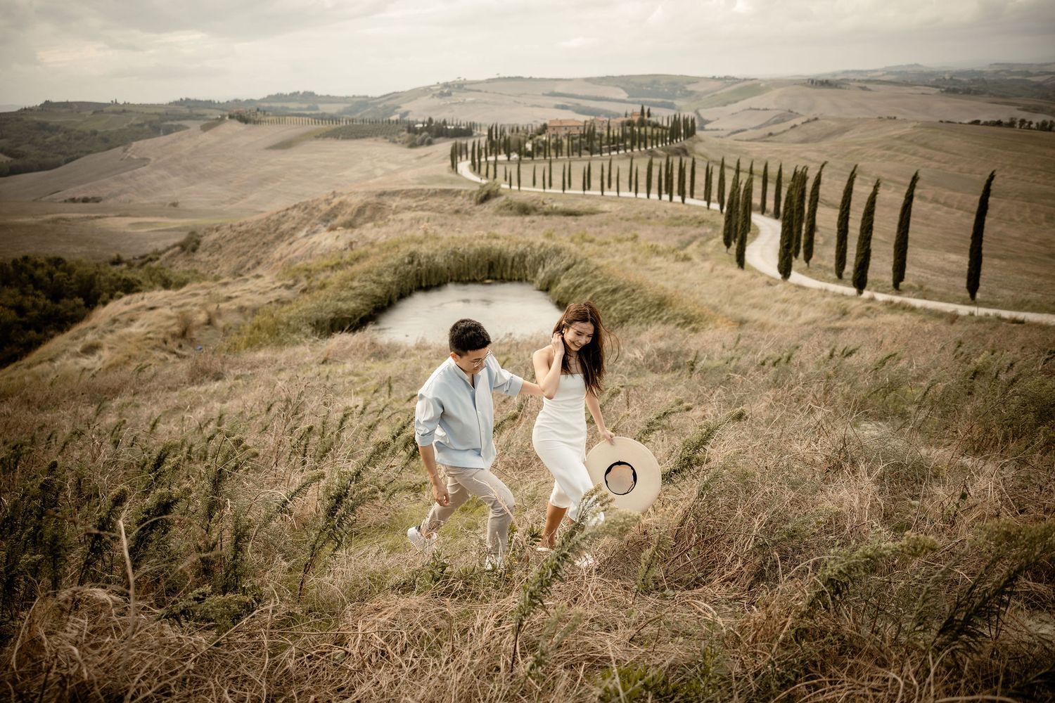Couple walking down scenic hillside path lined with towering cypress trees in Italian countryside.