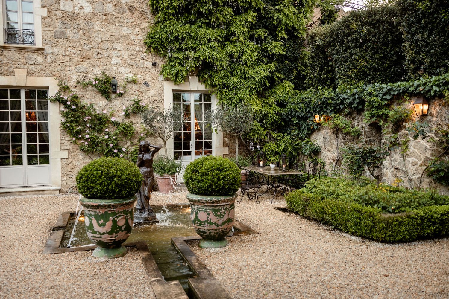 Charming stone courtyard with potted boxwoods and climbing ivy features gravel paths and French doors in rustic setting.