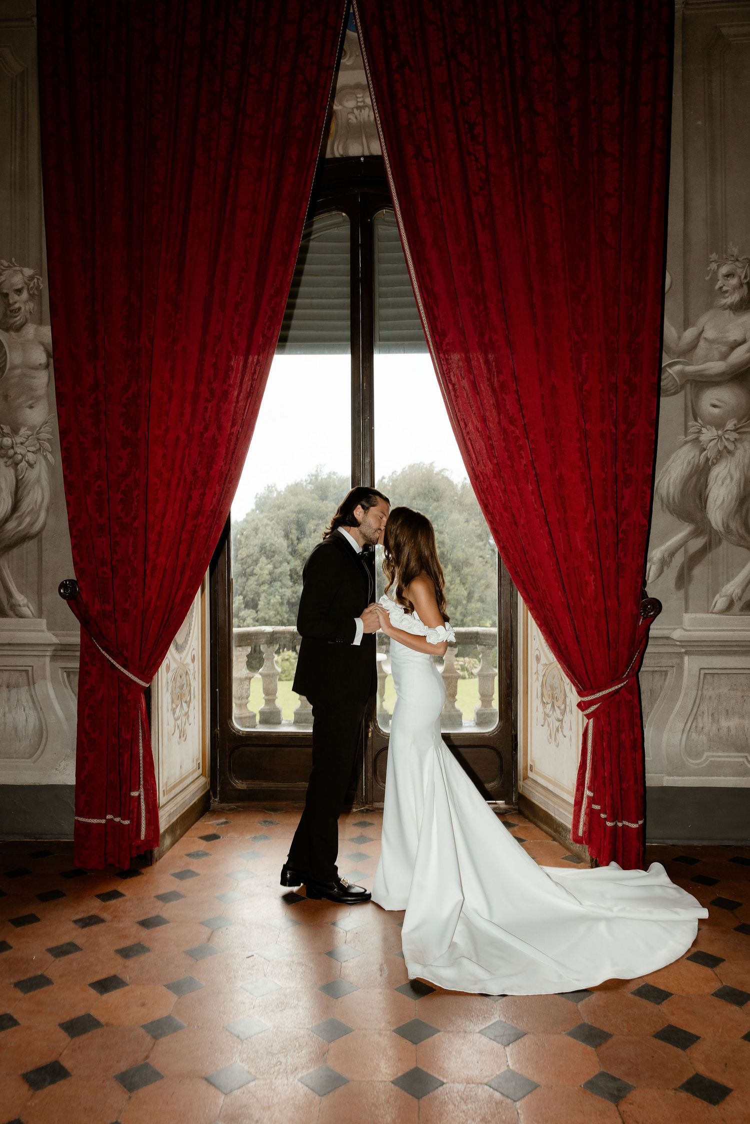 A couple shares a romantic moment between elegant red curtains and marble columns in a grand palace setting.