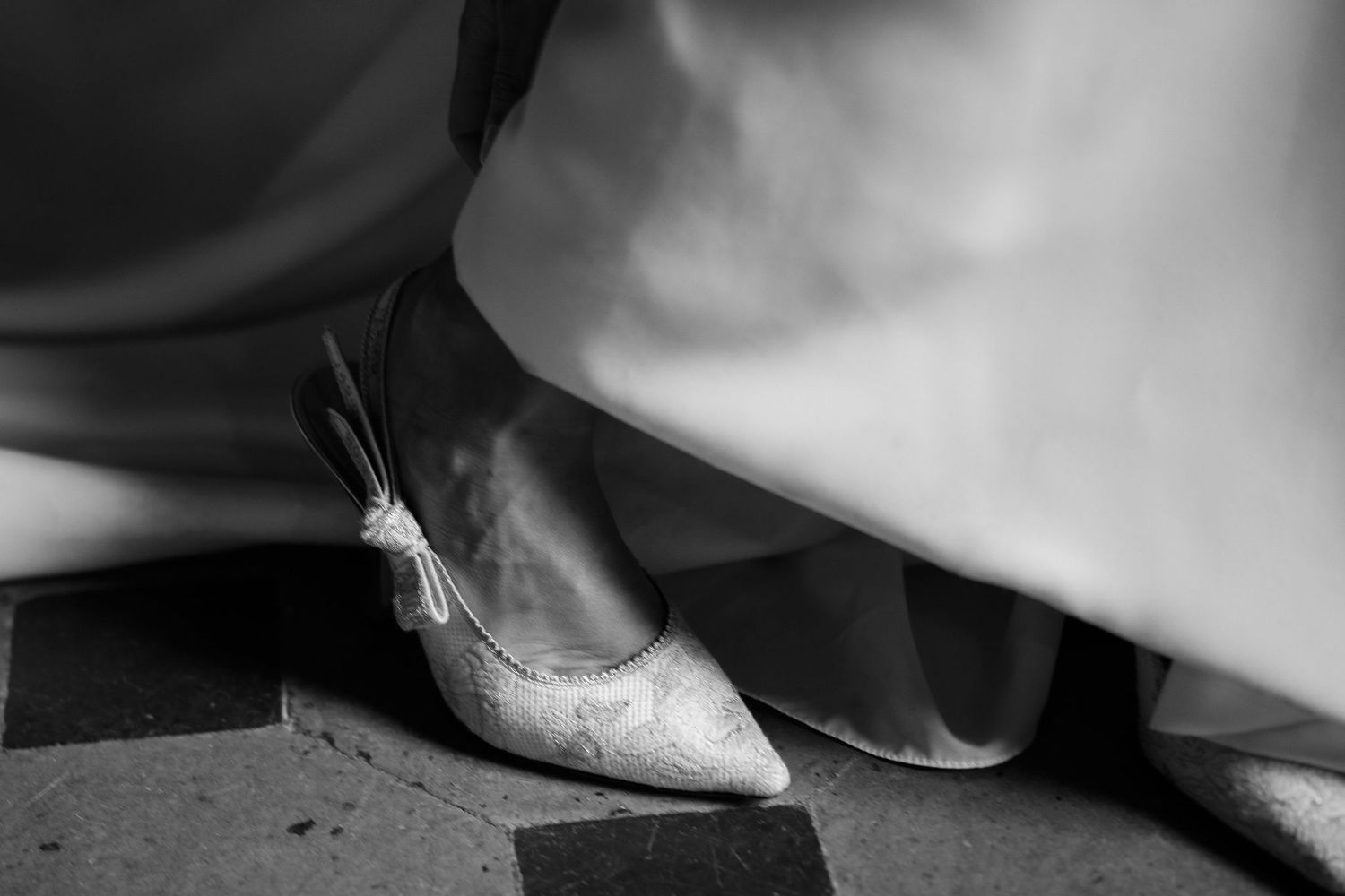 Black and white close-up photograph of an elegant pointed ballet flat shoe peeking out from under flowing fabric.