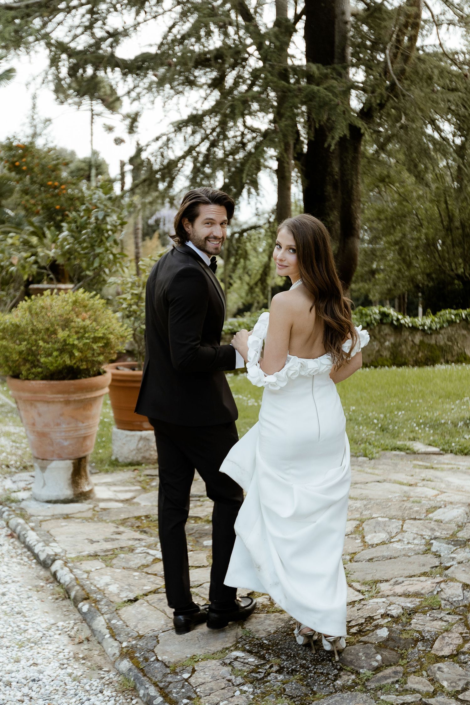 A bride in a white gown and groom in a black suit share a playful moment on stone steps in a garden setting.