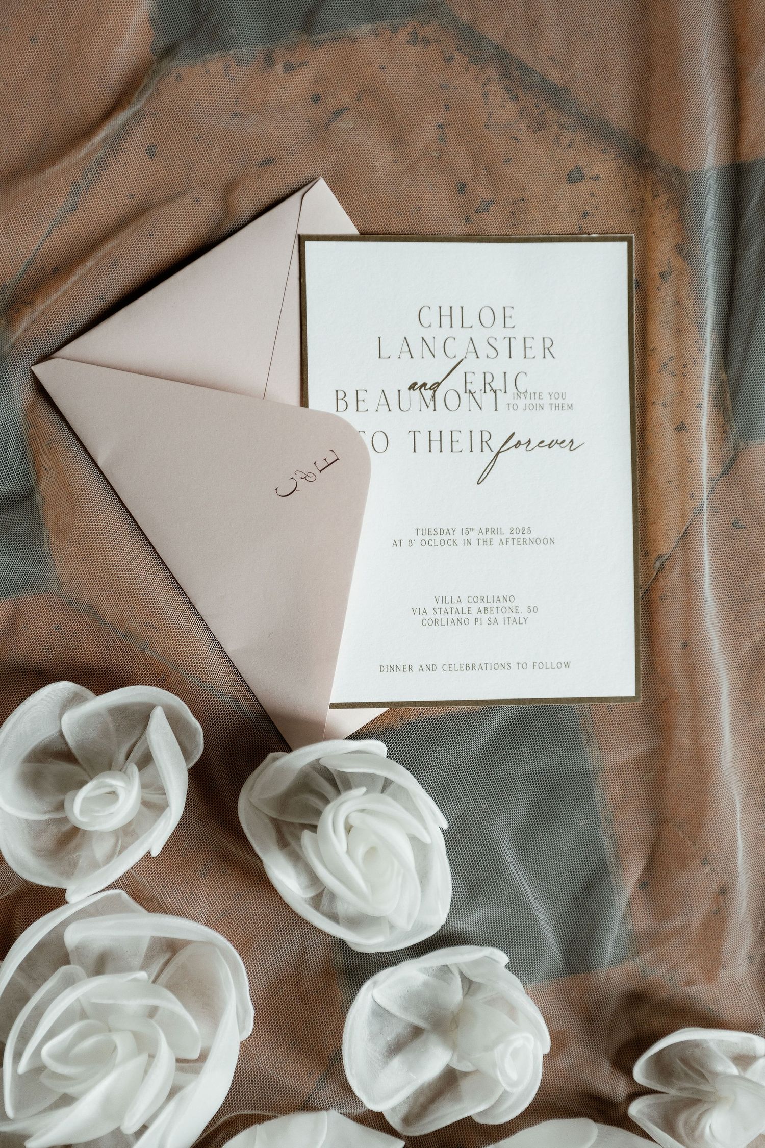 Elegant wedding invitation with white rose decorations and blush envelope against a neutral fabric background.