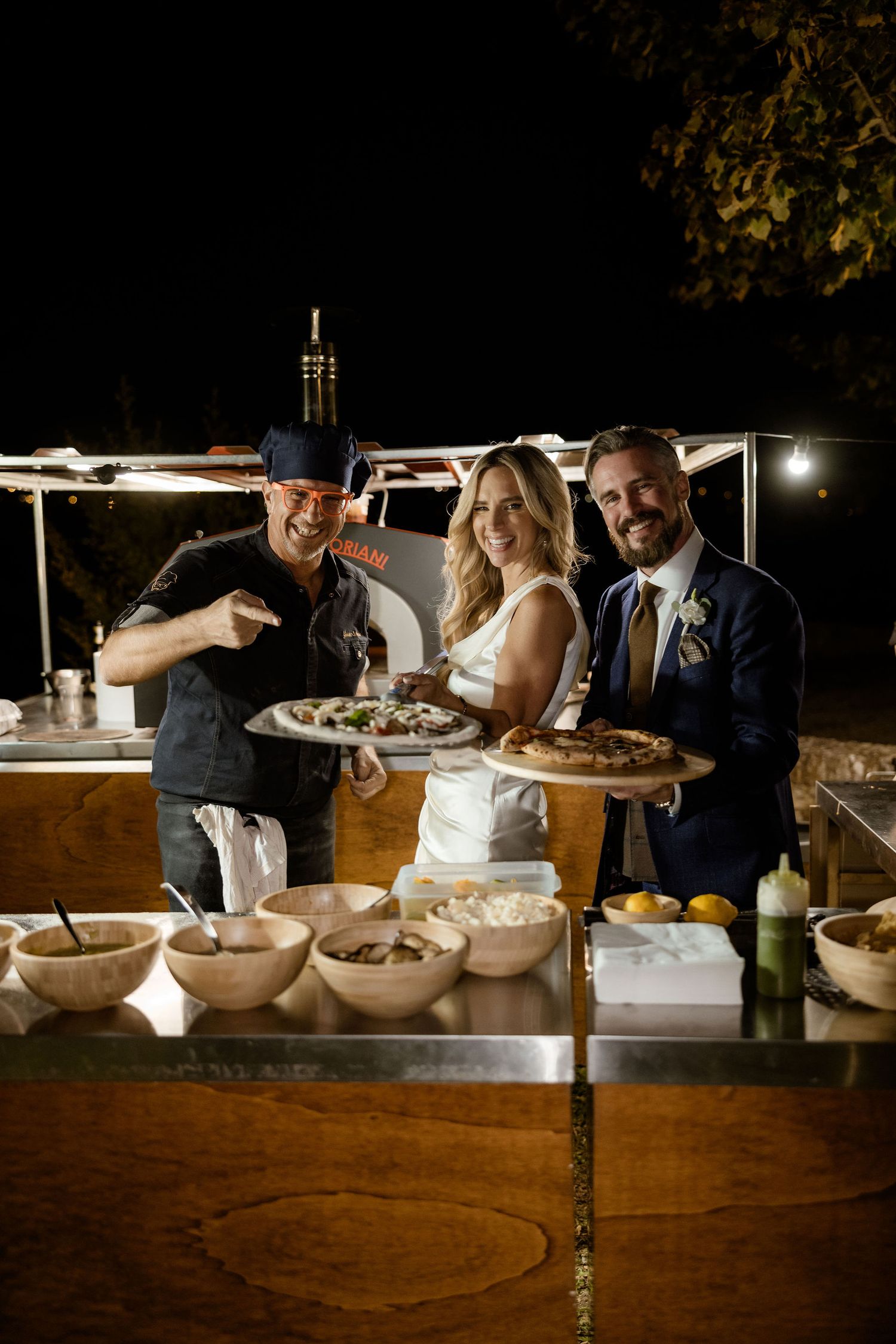 Guests enjoying food and drinks at a rustic wooden buffet table during an evening outdoor event.