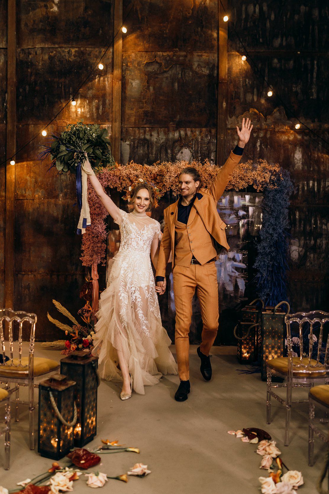 Couple celebrates joyfully in rustic venue with warm lighting and scattered rose petals on concrete floor.