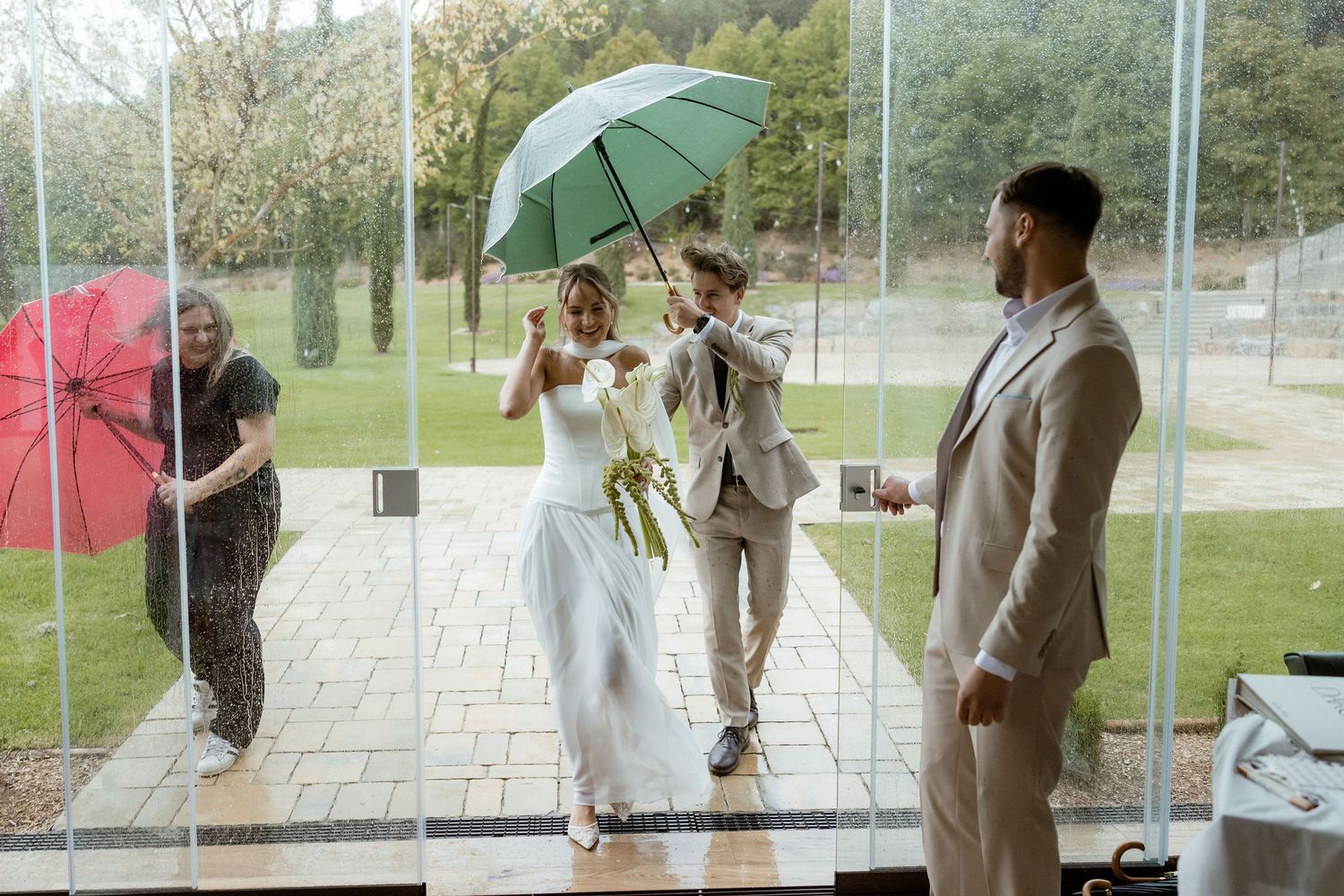 Wedding party members pose with colorful umbrellas on a rainy day outside during a celebration.