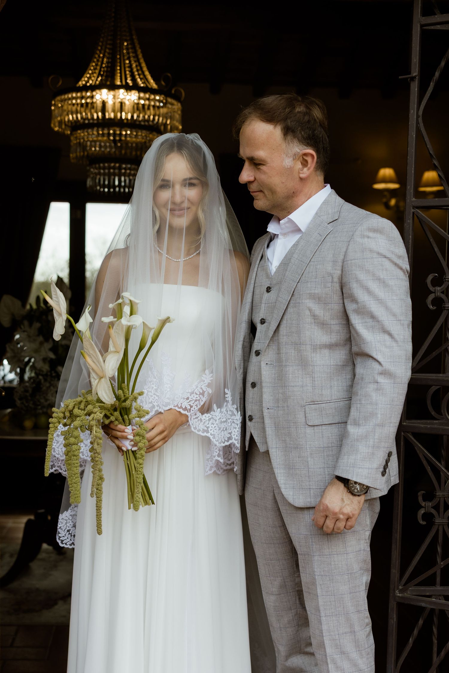 A wedding couple stands together, with the bride in white lace dress and groom in light gray plaid suit at their ceremony.