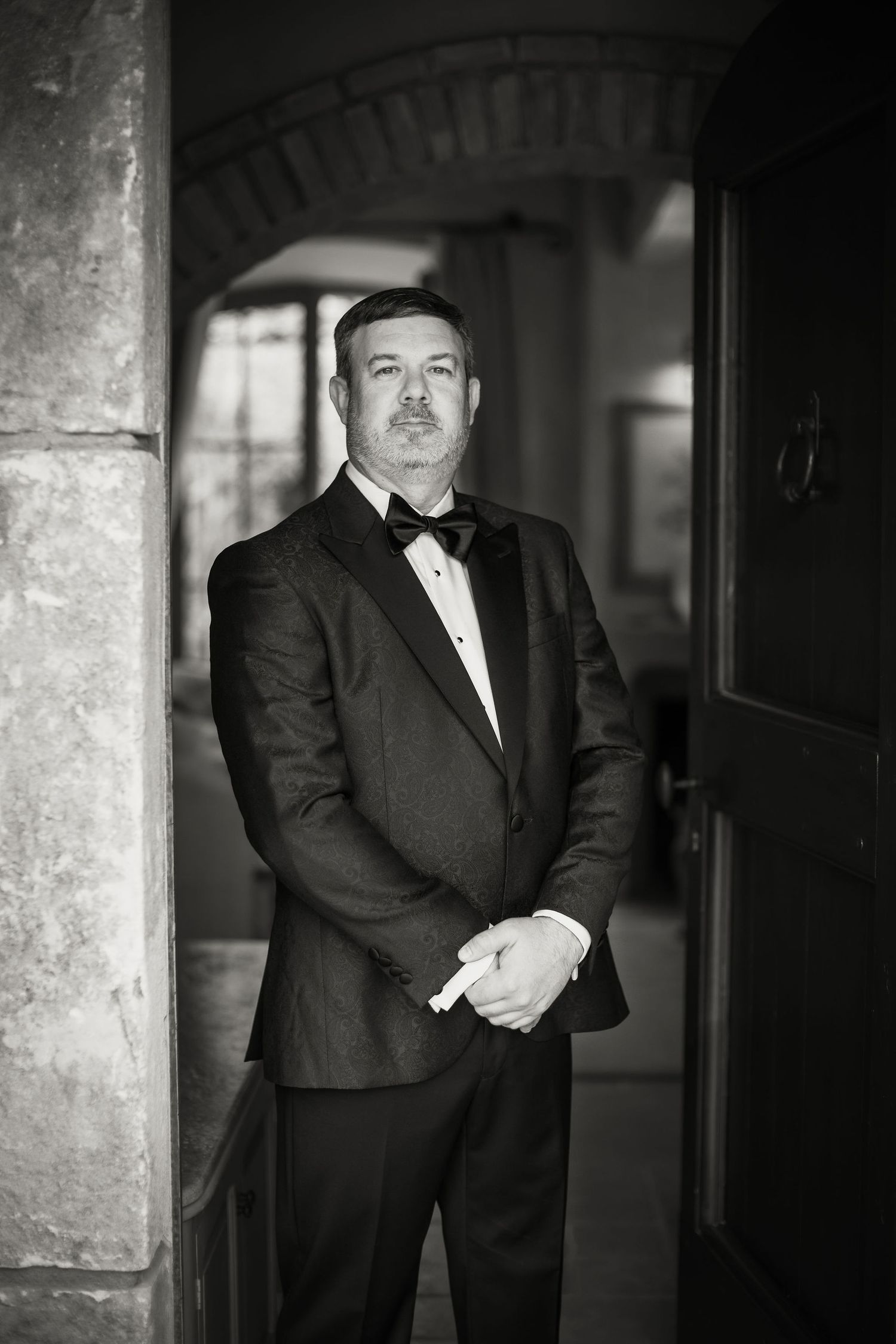 Black and white formal portrait of someone in a tuxedo standing in an arched doorway.