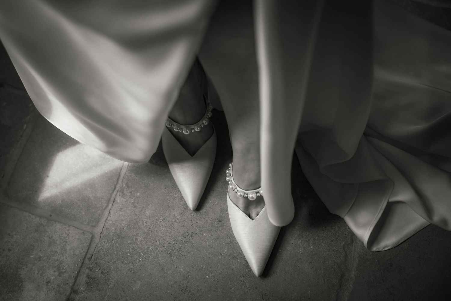 Black and white wedding photo showing elegant white high heels and groom's dress shoes on stone floor.