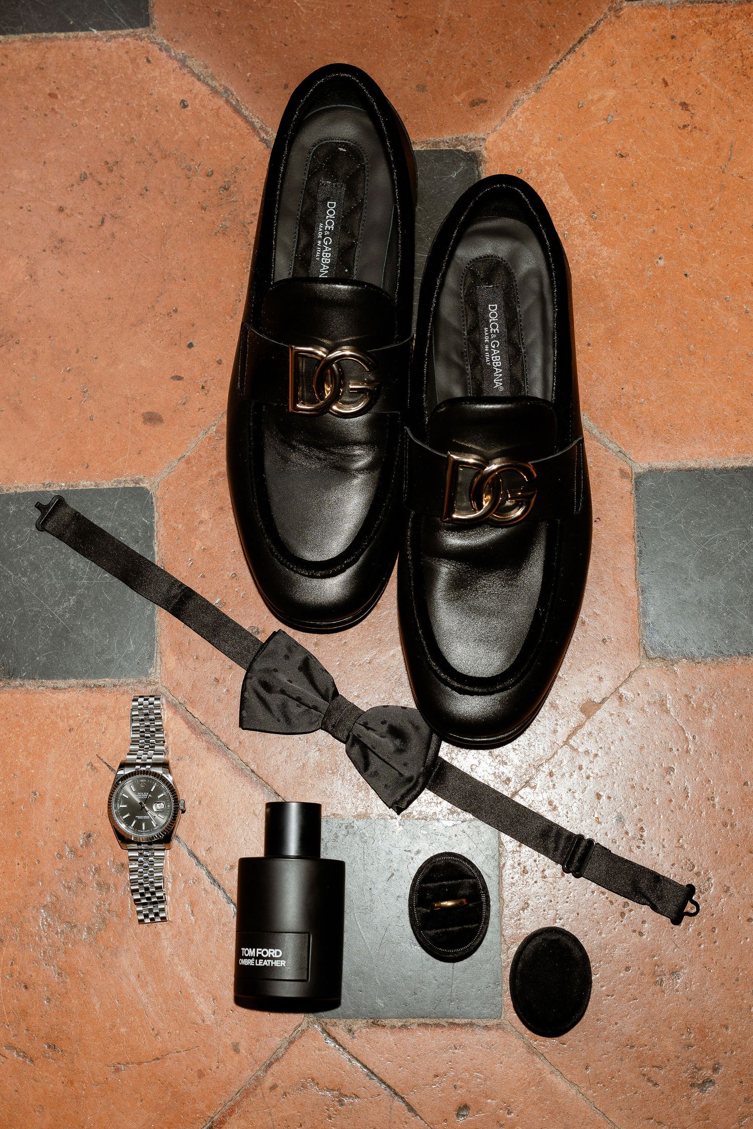 Black leather loafers, bowtie, watch and fragrance bottle arranged on terracotta tile floor in monochromatic styling.