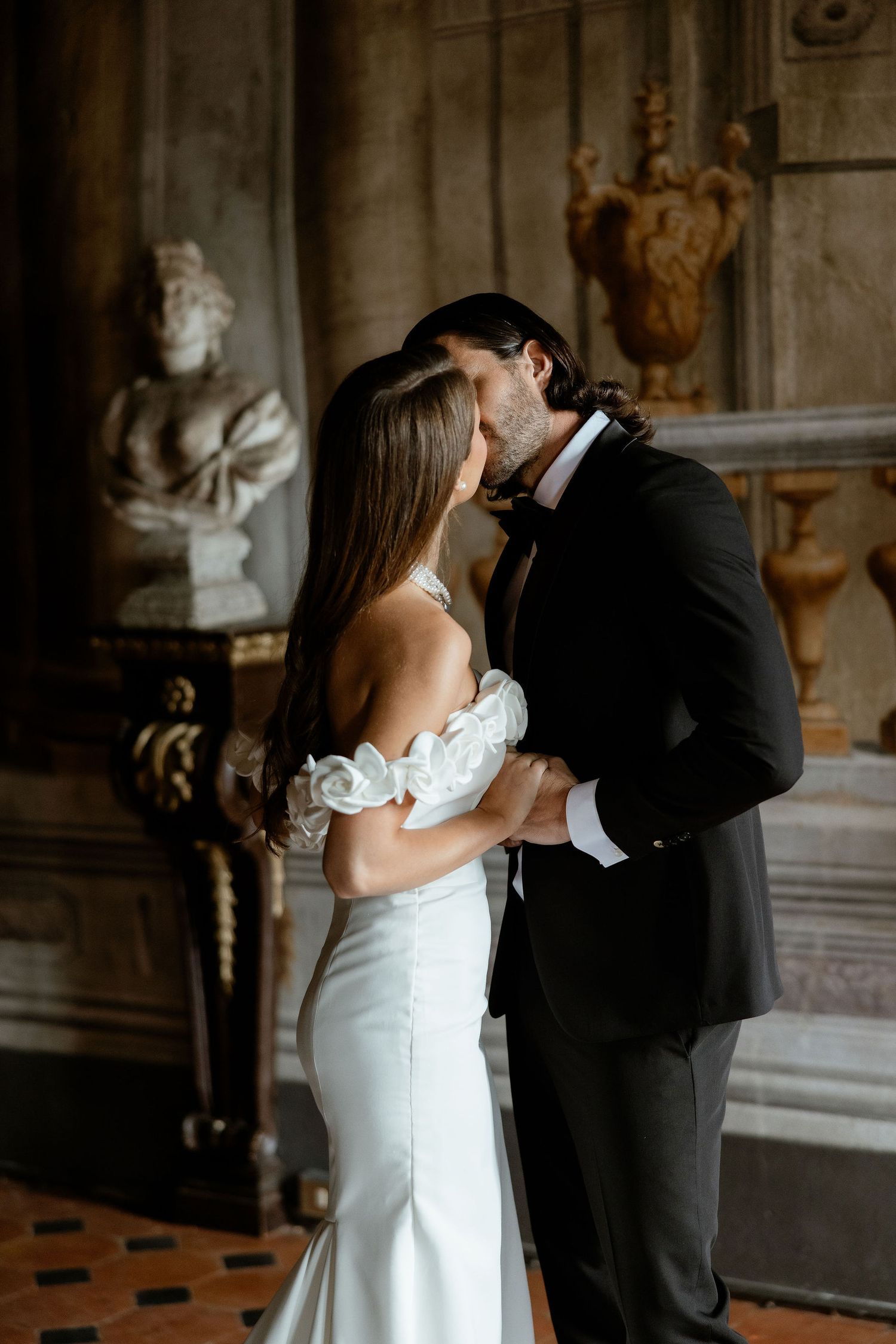 Couple shares intimate moment during wedding photoshoot in ornate historical building with classical sculpture backdrop.