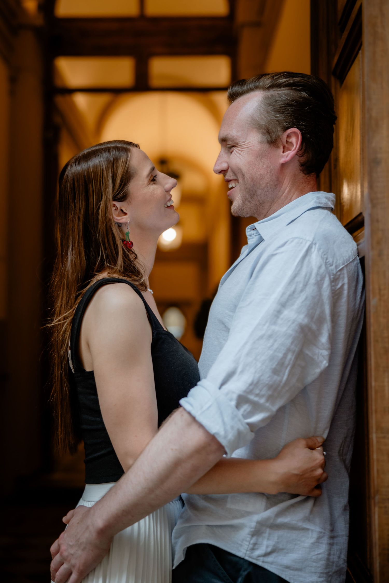 A romantic couple shares an intimate moment in an elegant hallway with warm lighting and arched architecture.