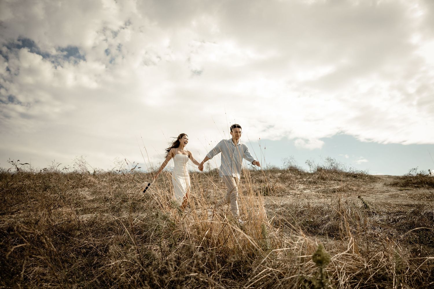 Couple walking hand in hand through dry grass field under cloudy sky.