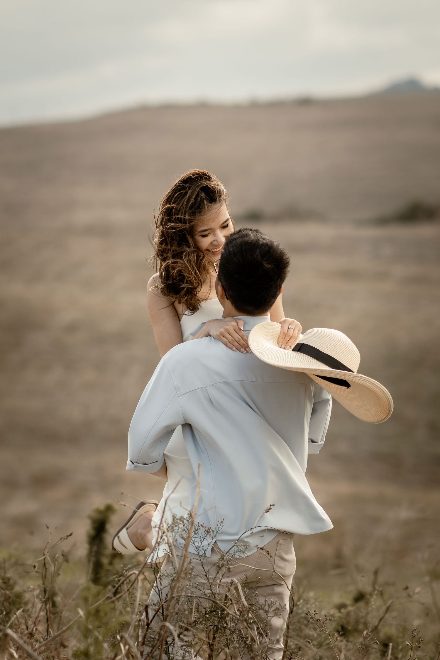 A romantic couple embraces on a hilltop against a moody desert landscape at sunset.
