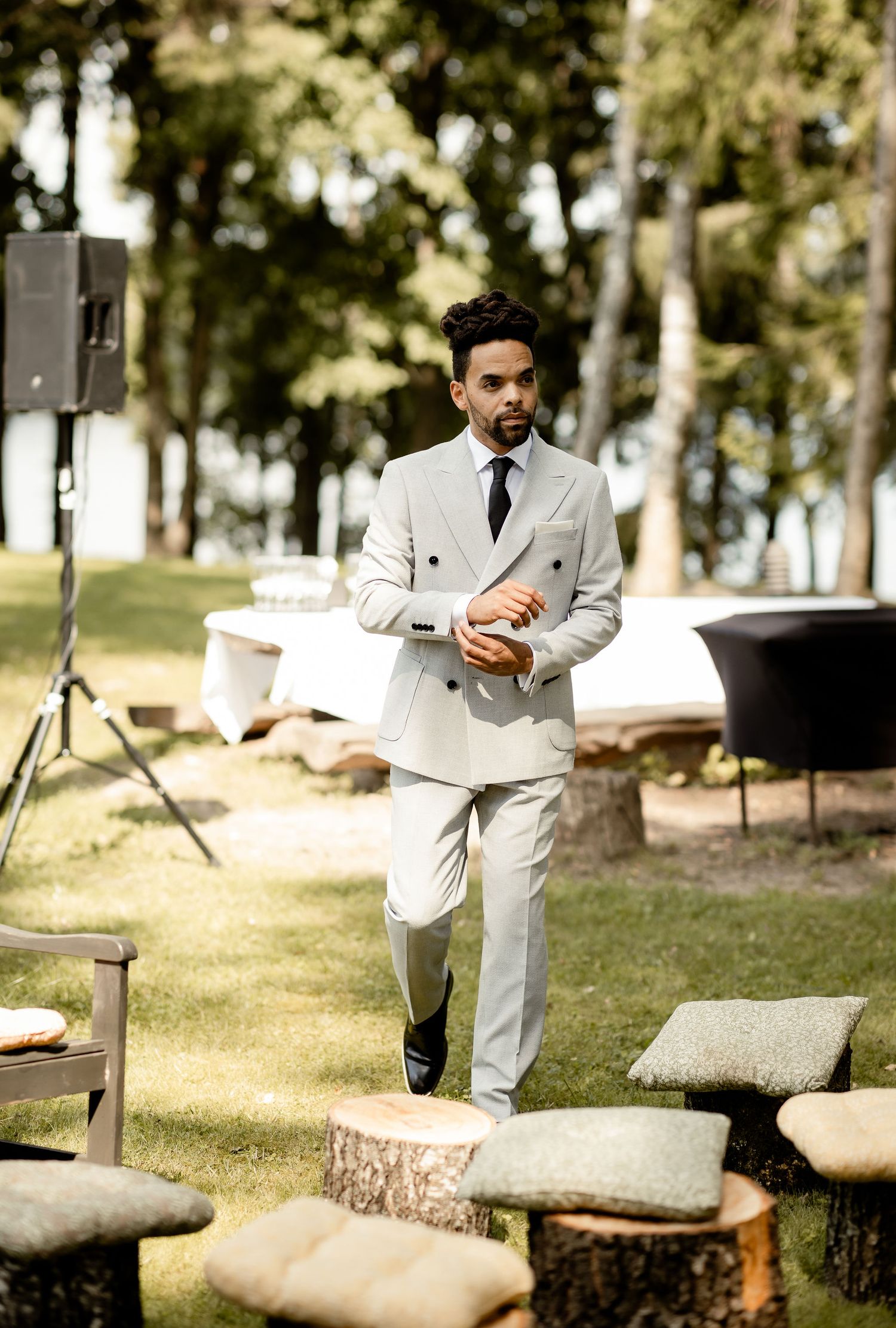 Stylish person in light grey suit walking across outdoor wedding setup with rustic elements.