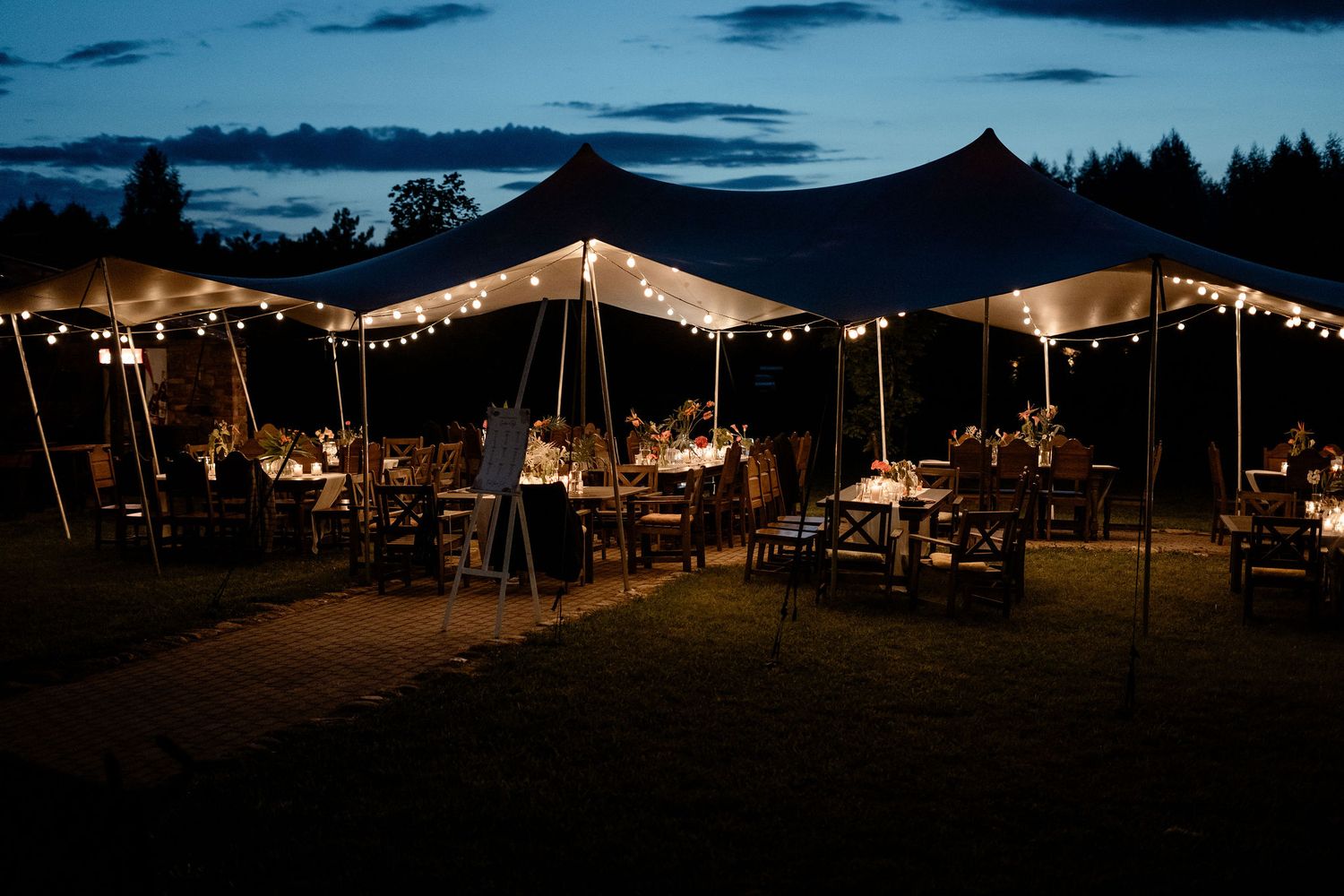 An outdoor wedding reception under illuminated tents with string lights at dusk, featuring tables and chairs on a lawn.