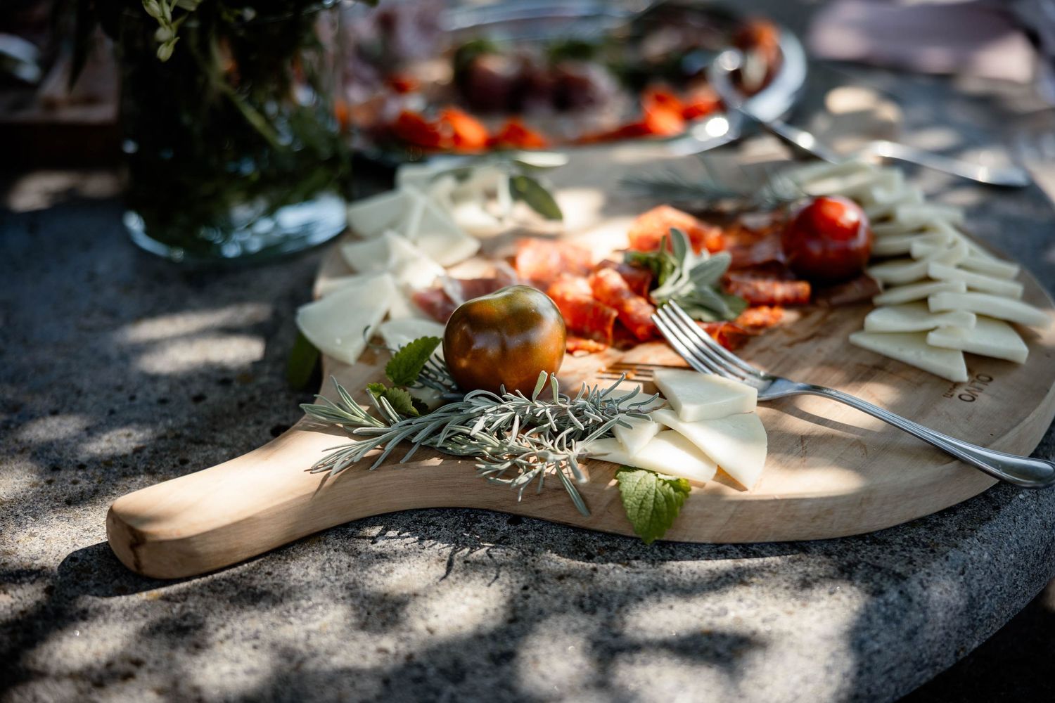Appetizers and fresh herbs arranged on a wooden serving board with dappled sunlight.