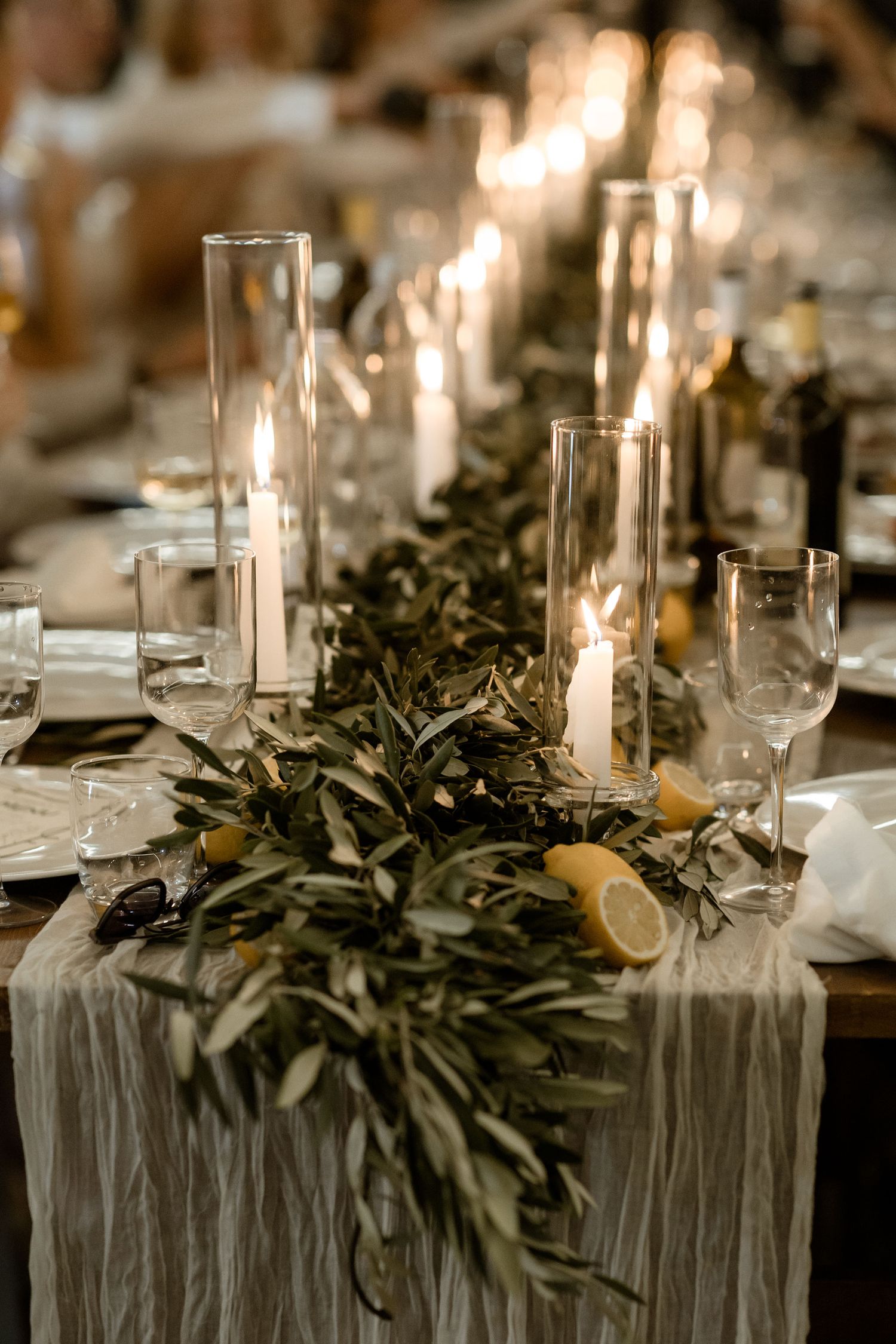 Elegant wedding table setting with tall glass candle holders and olive branch garland on gray linen runner.
