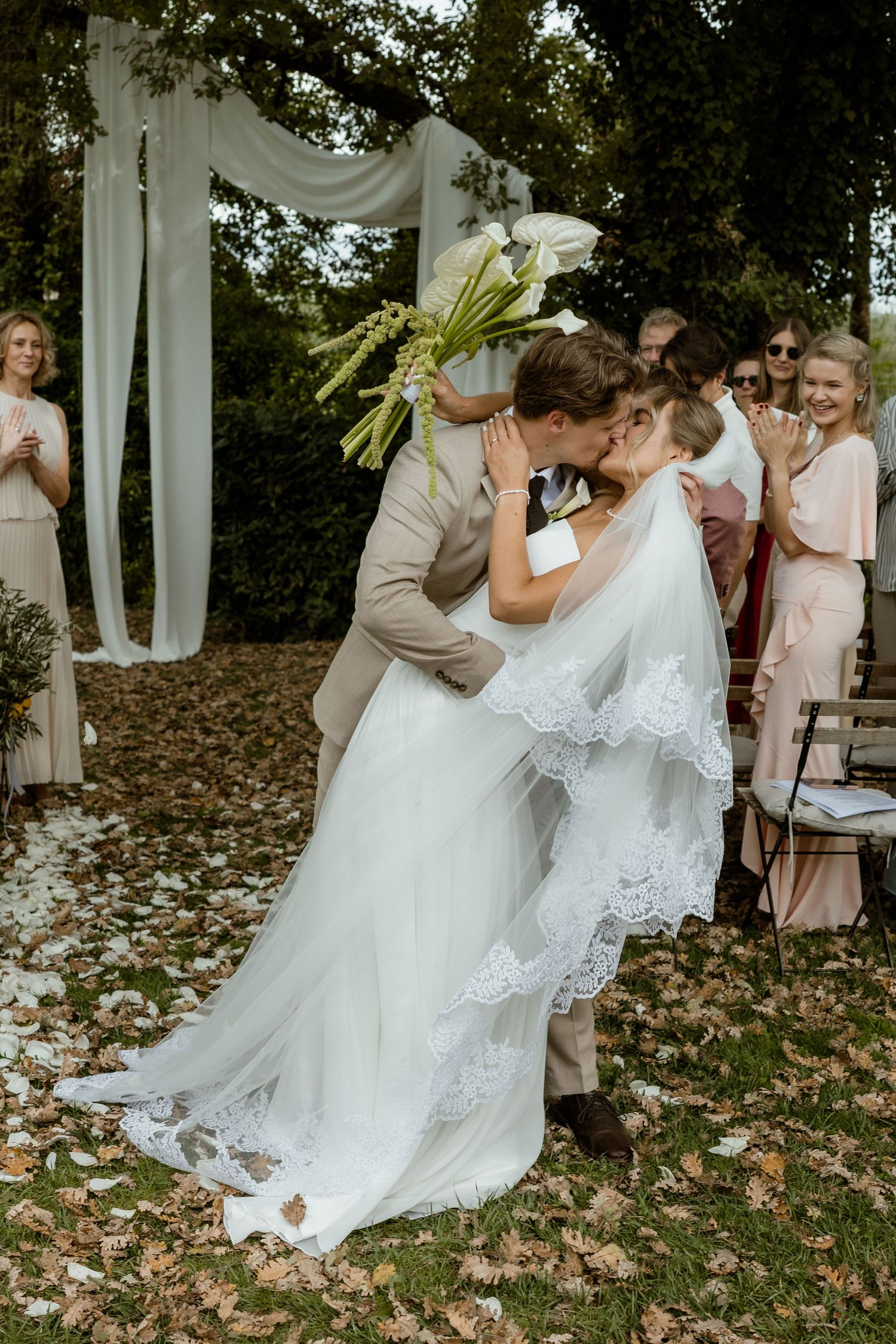A romantic outdoor wedding ceremony with a white draped arch, autumn leaves on the ground, and bridesmaids in blush dresses.