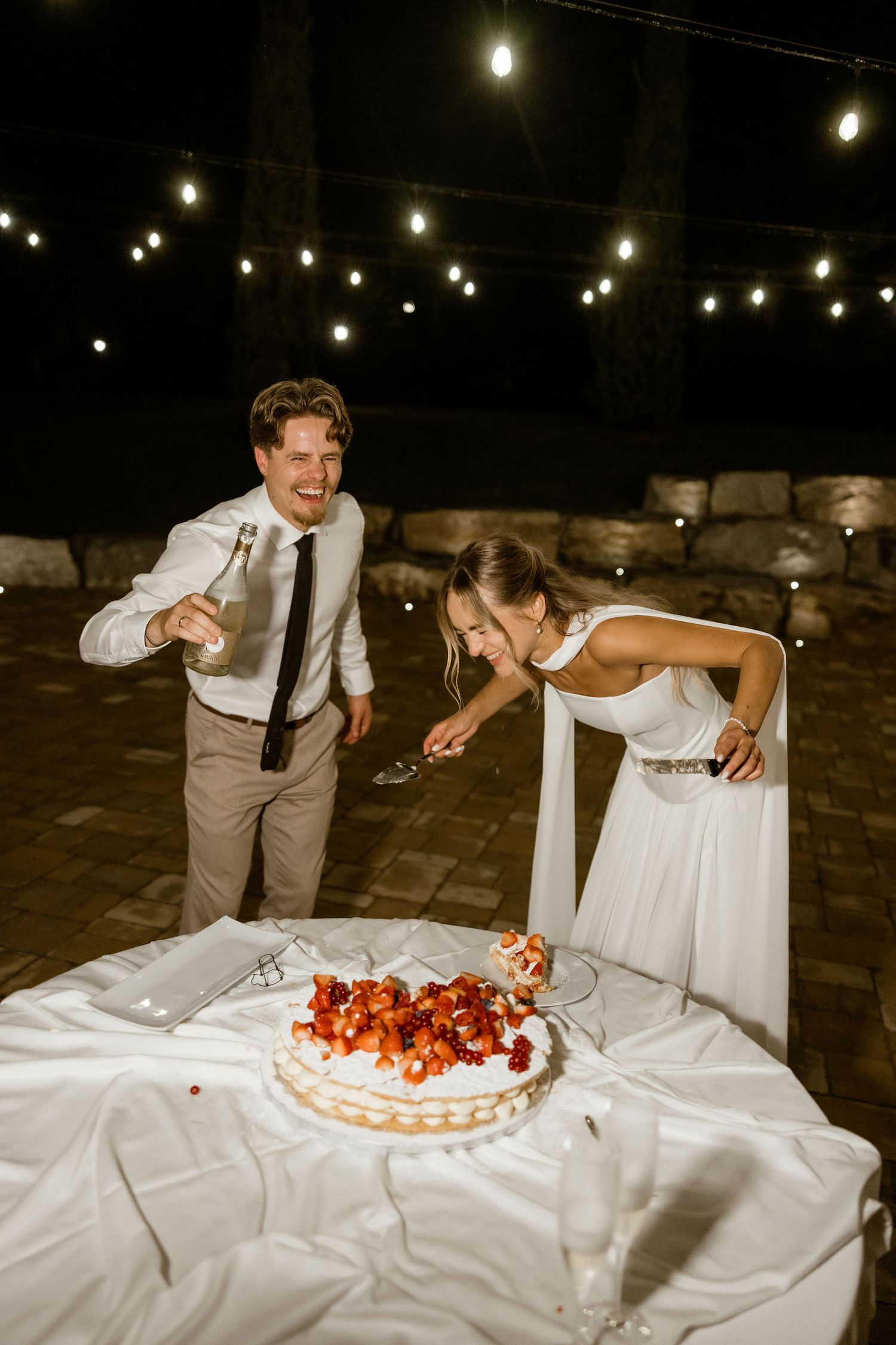 Wedding couple playfully smashes cake together during reception under string lights at night.