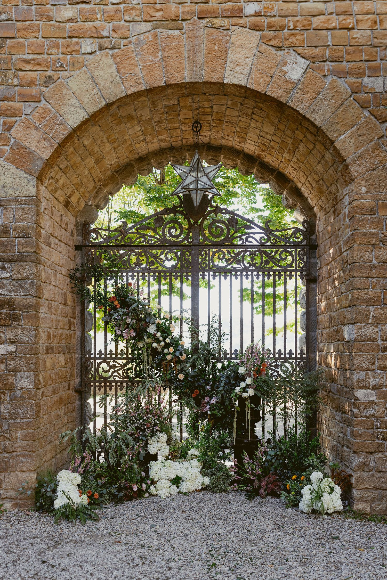Ornate wrought iron gate with white flowers beneath a brick archway in an Italian garden setting.