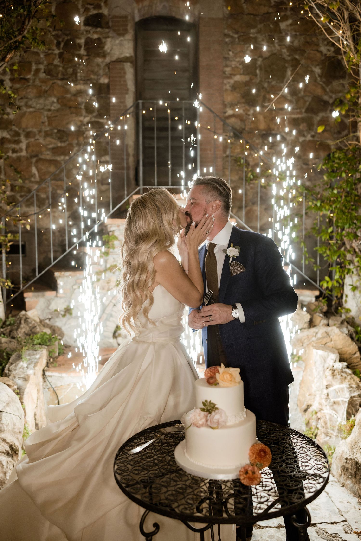 A romantic wedding moment with sparklers and a white tiered cake on a decorative stand at a rustic stone venue.