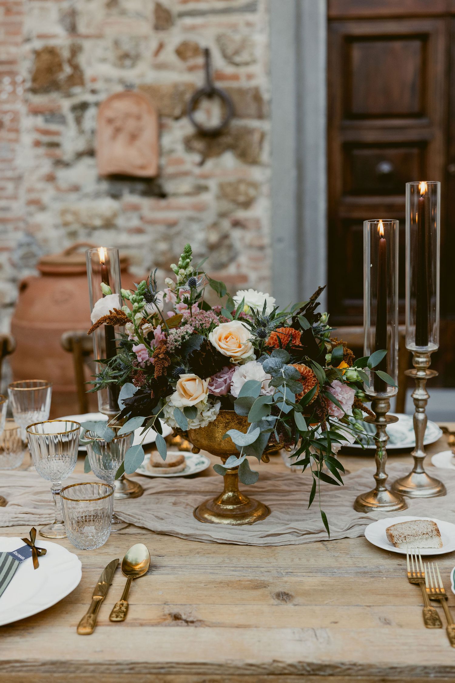 Elegant rustic wedding table setting with floral centerpieces, brass candlesticks, and vintage place settings against stone wall.