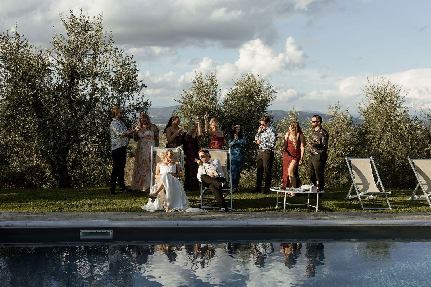 Wedding party stands near a reflective pond surrounded by trees on a cloudy day for outdoor photo session.