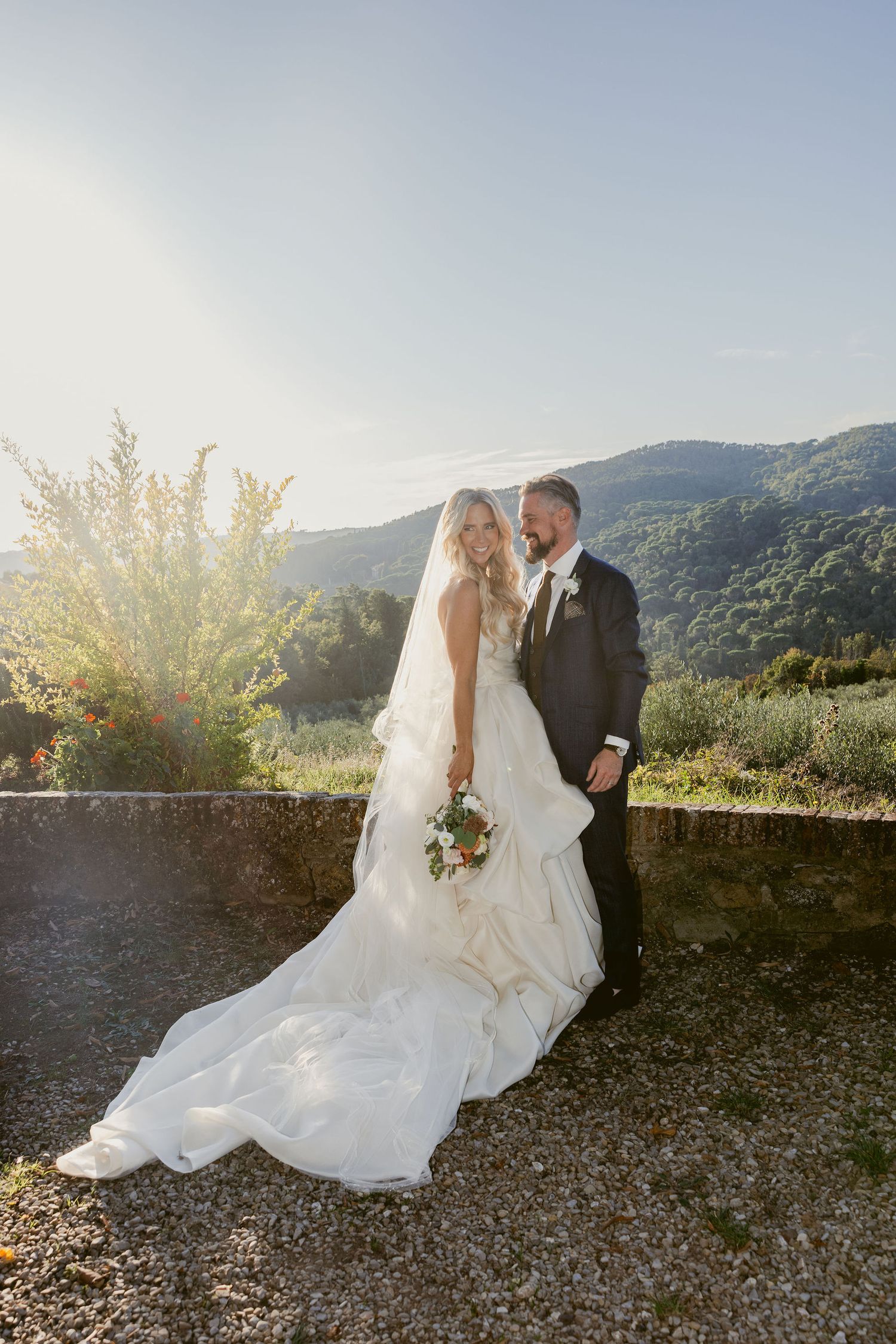 A romantic wedding couple poses together against a scenic mountain backdrop at sunset with flowing white dress.