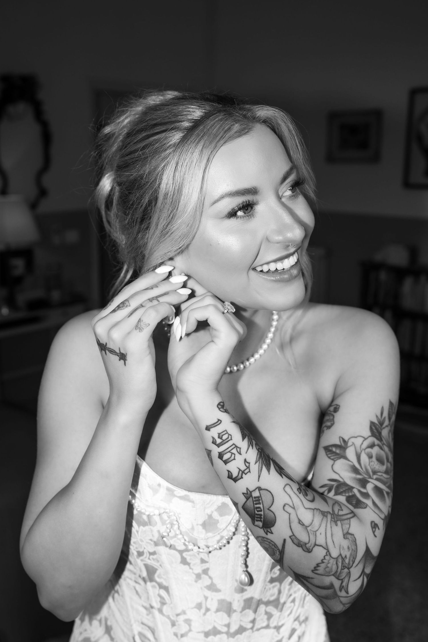 Black and white portrait of a smiling bride in lace dress with pearl necklace and visible arm tattoos.