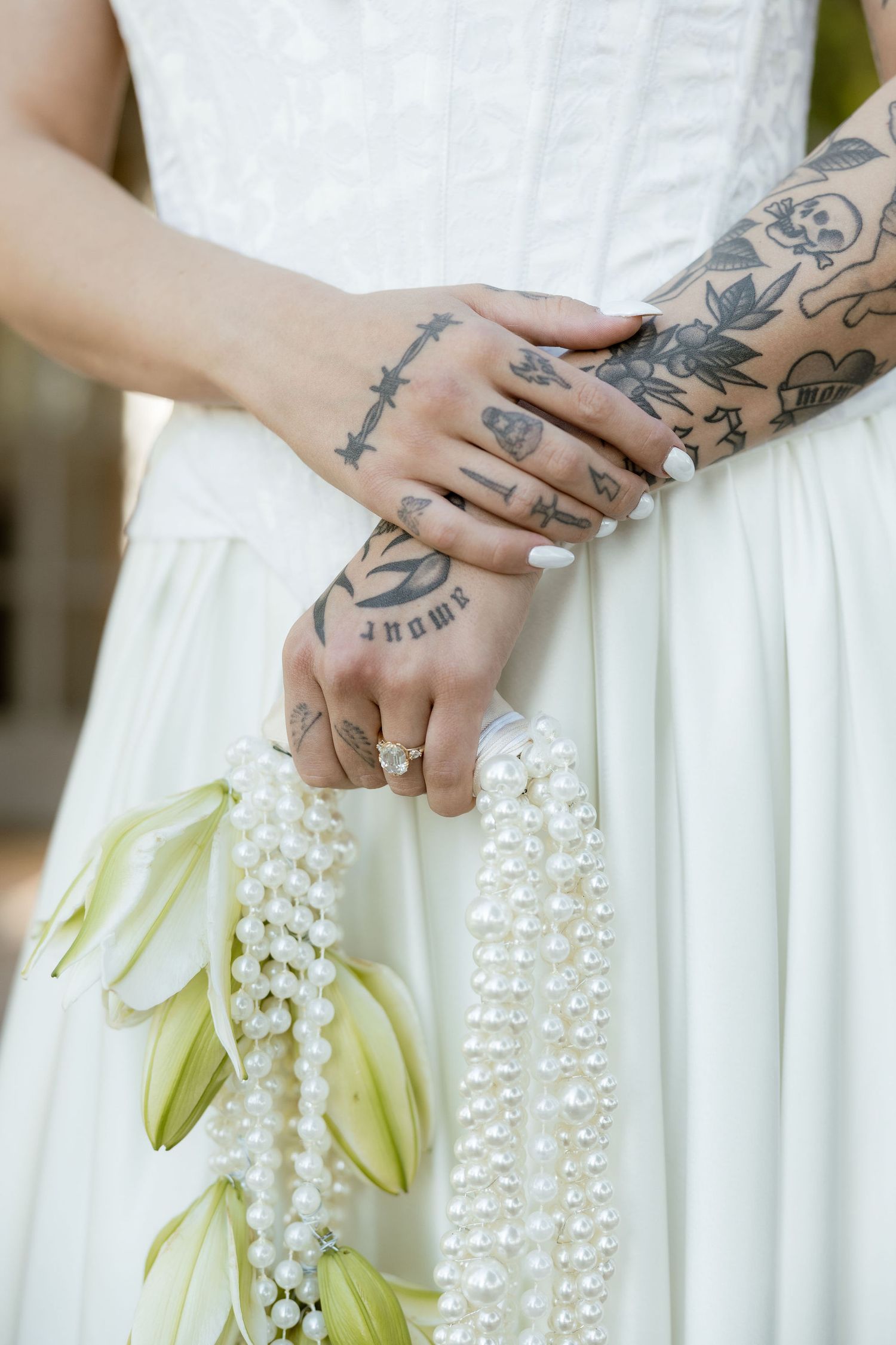 Close-up of tattooed hands holding white lilies and pearl strands against a flowing white dress.