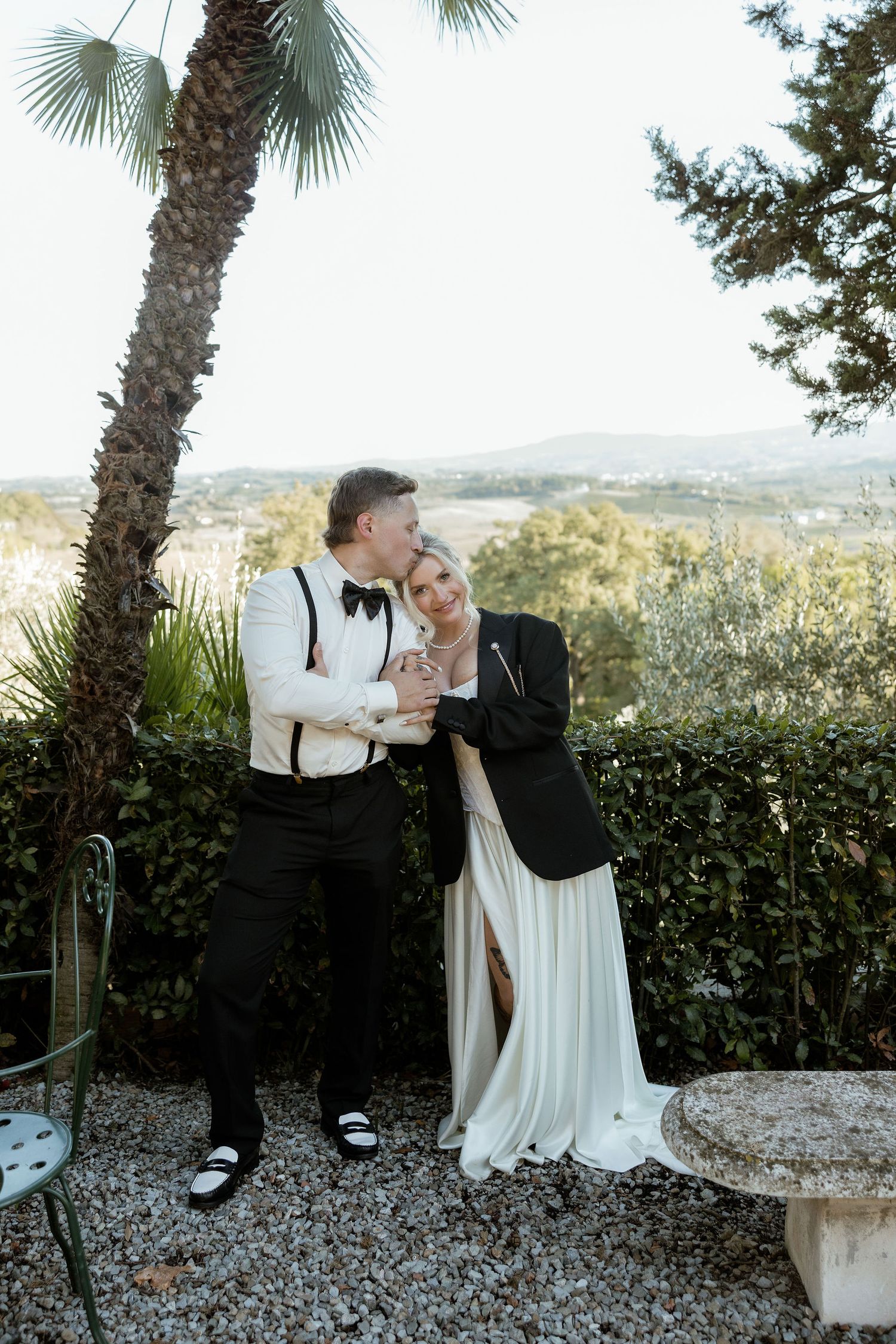A romantic wedding couple embraces under palm trees with a scenic hillside view in the background.