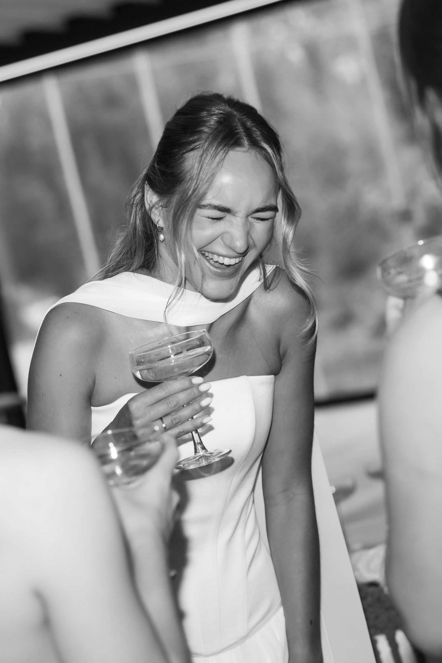 Black and white candid photo of someone in a white dress laughing while holding a cocktail glass at an event.
