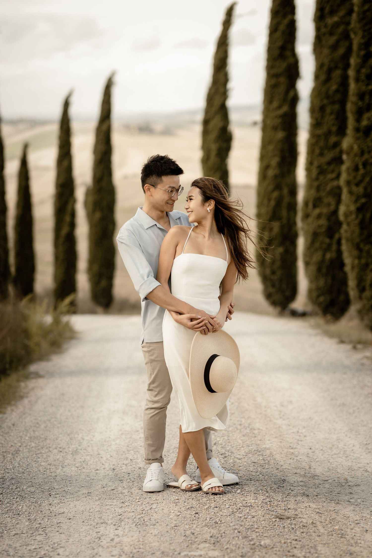 Couple sharing tender embrace while holding straw hat on scenic Italian cypress tree road.