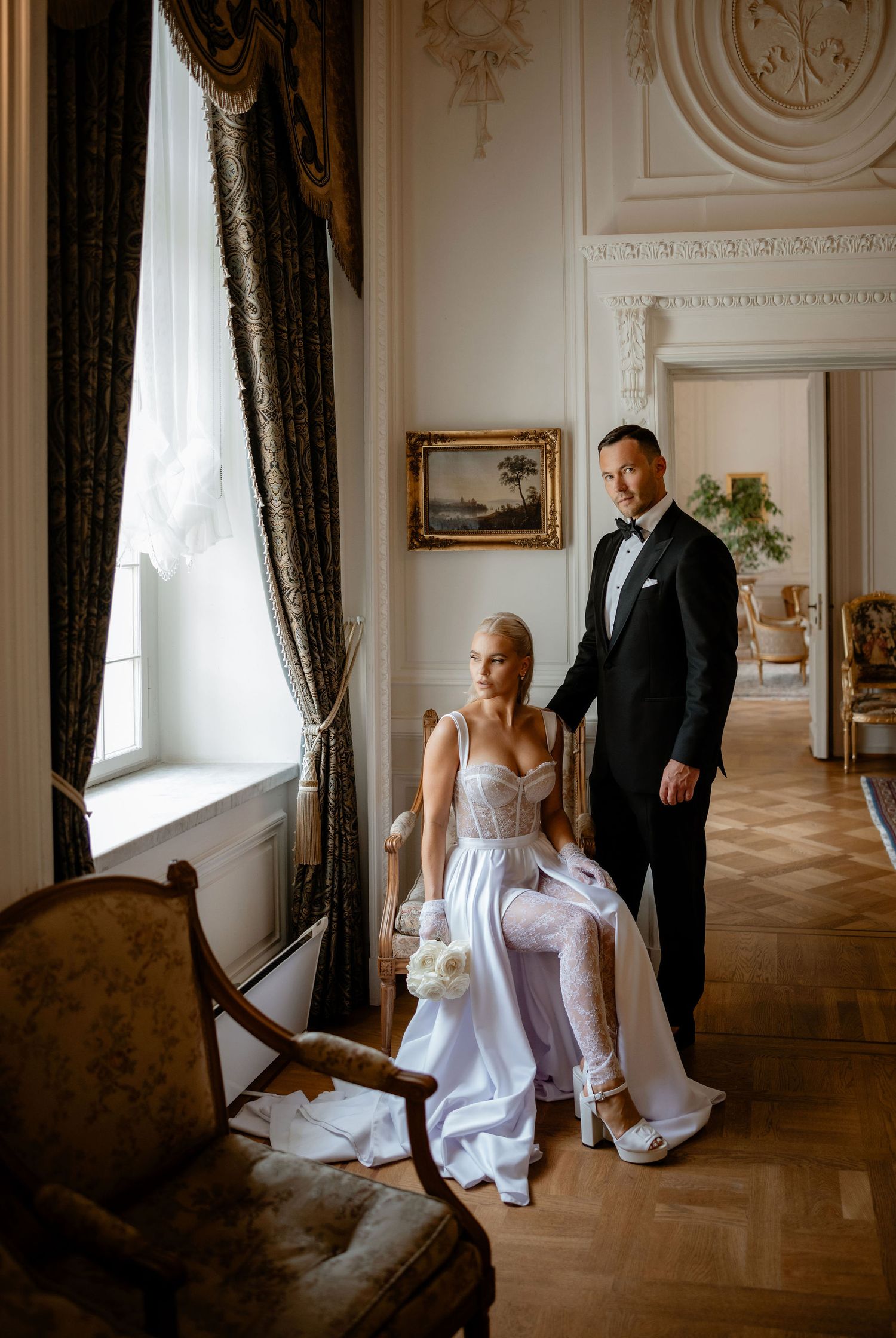 Bride and groom share an intimate moment in an ornate room with antique furniture and flowing curtains.