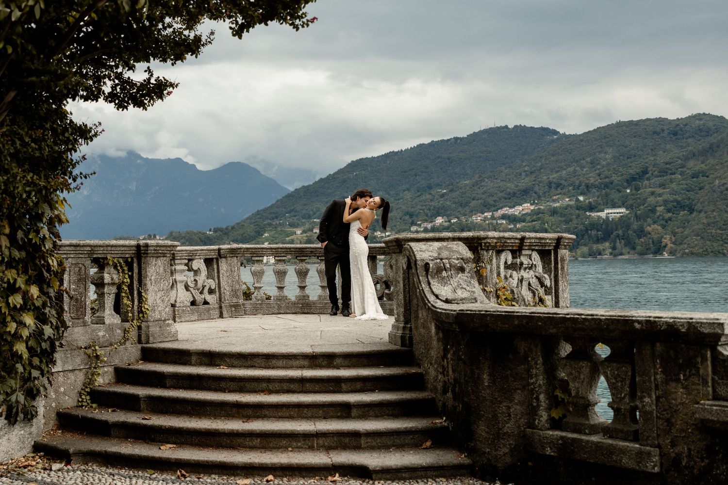 Wedding couple on stone steps of ornate terrace overlooking mountain lake landscape.